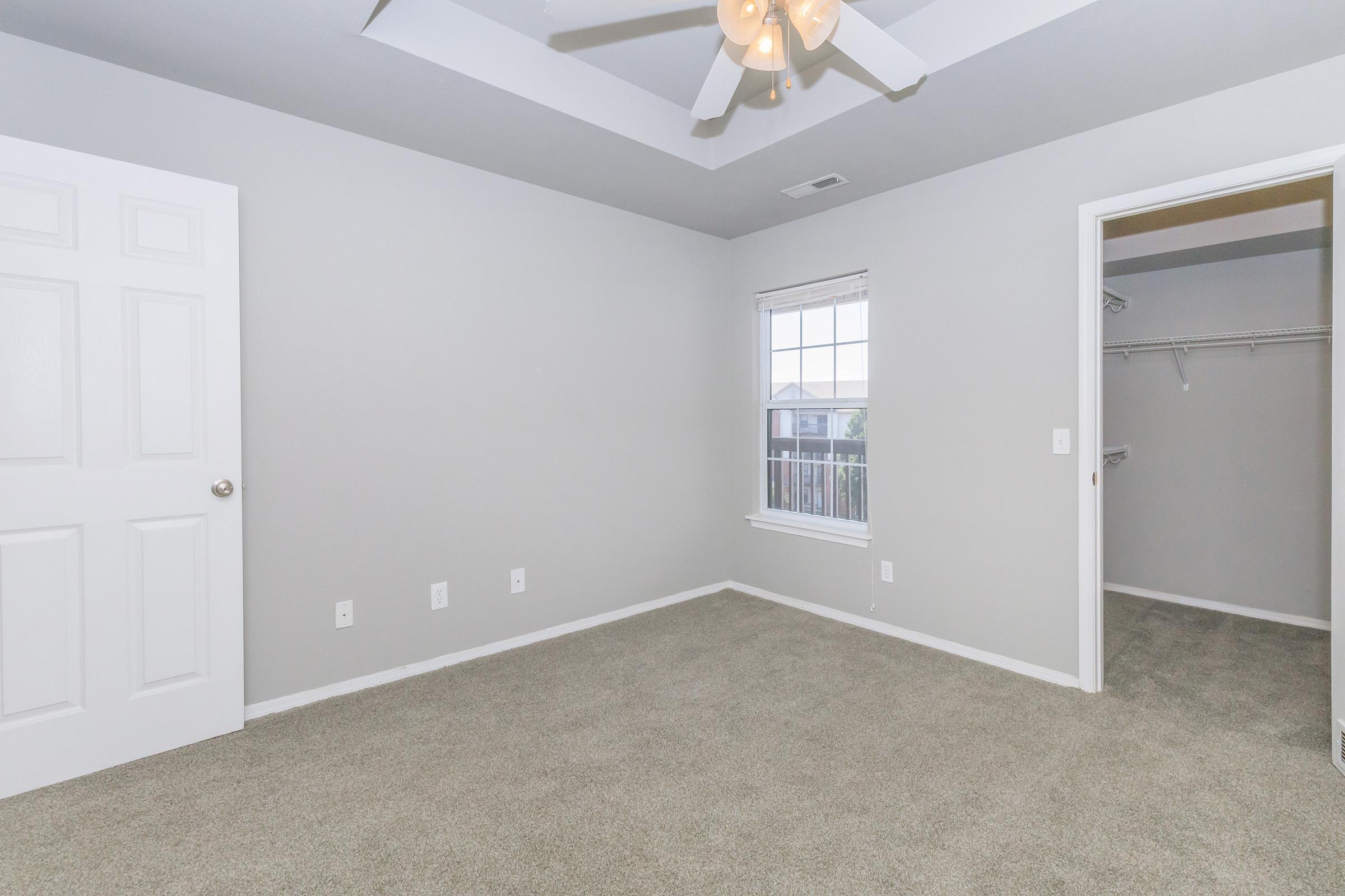 Empty bedroom featuring light gray walls and carpet, a ceiling fan with lights, a window with white trim, and a closet with sliding doors. The room has minimal furnishings, emphasizing a spacious and bright environment.