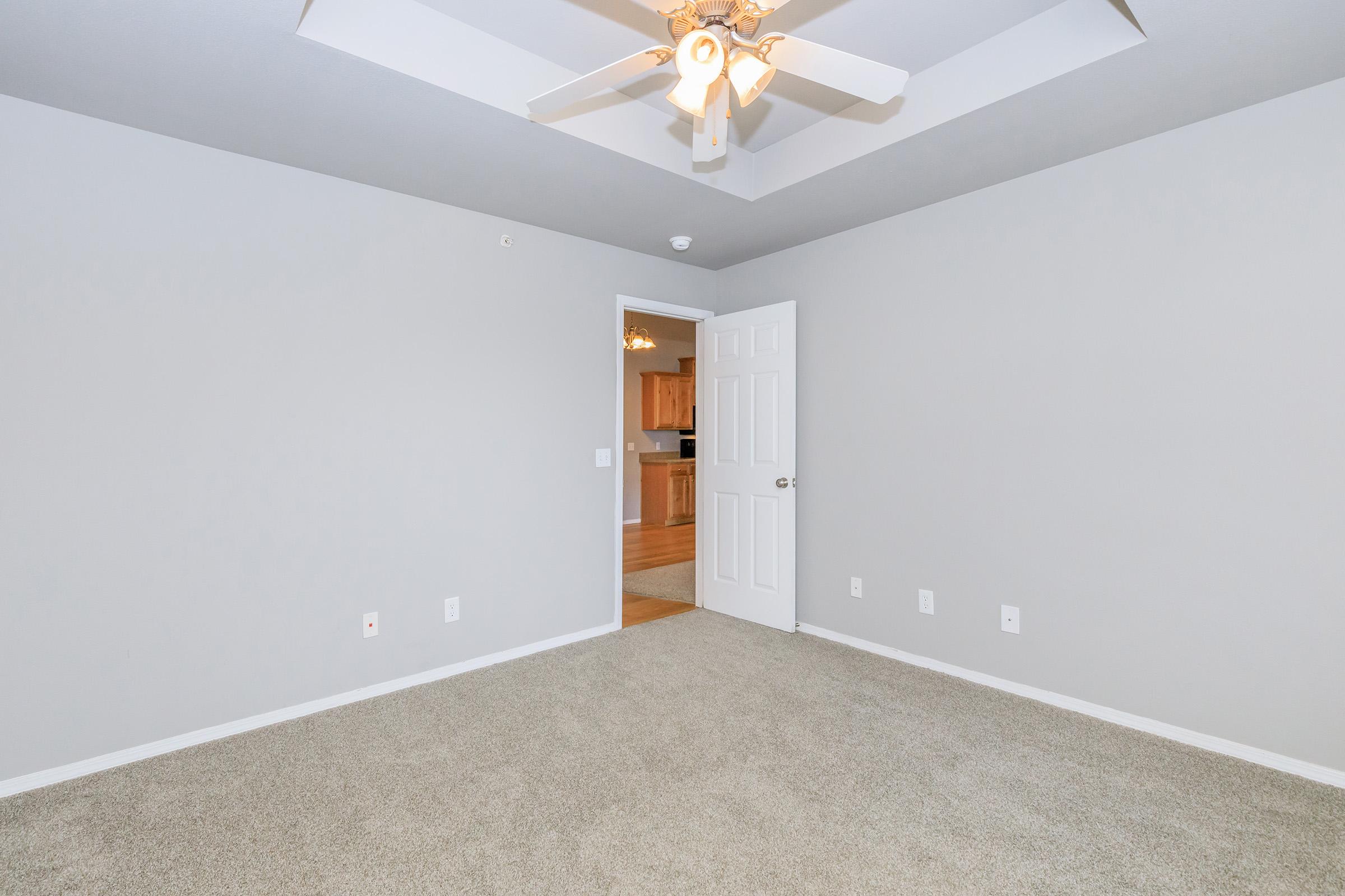 A well-lit empty room featuring beige carpet, light gray walls, and a ceiling fan. A doorway leads to an adjacent area, possibly a kitchen, visible in the background. The room is unfurnished and spacious, with three light switches on the wall.
