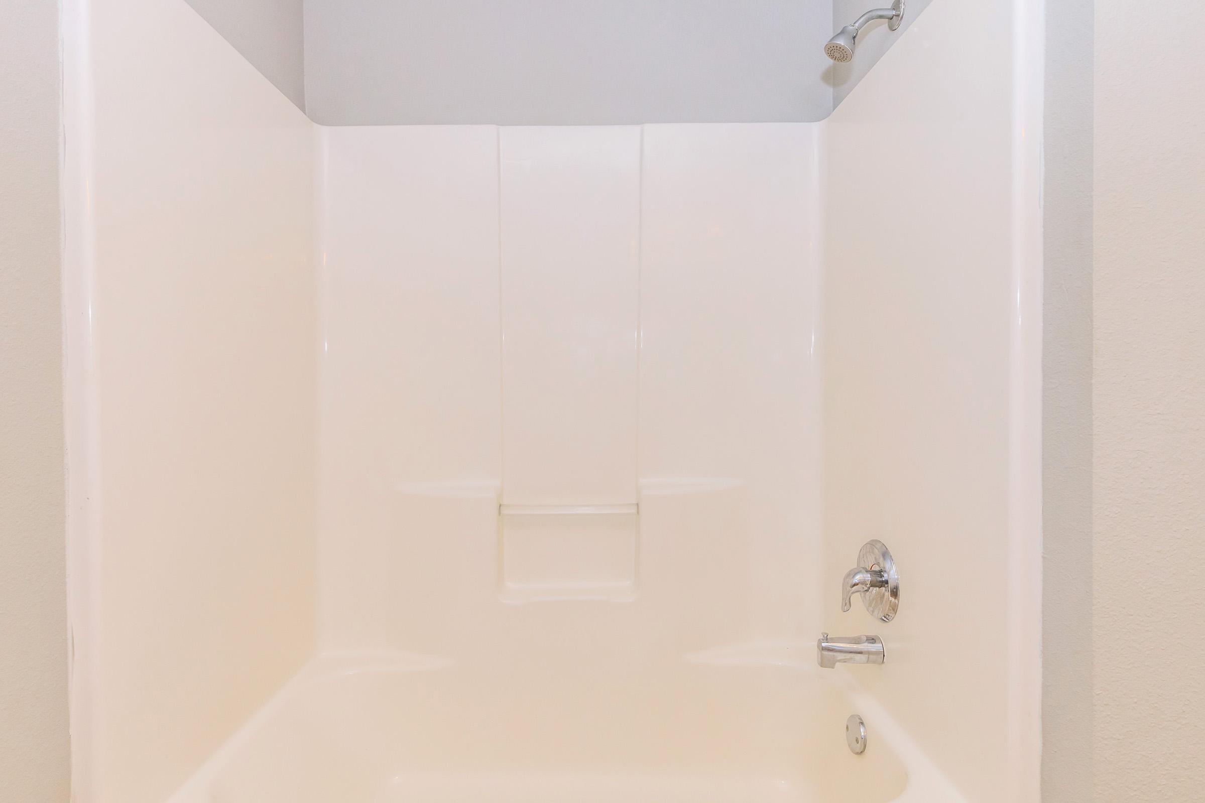 A clean, empty shower stall with smooth white walls, a showerhead, and a faucet visible. The stall features a low wall and is well-lit, creating a bright and spacious appearance.