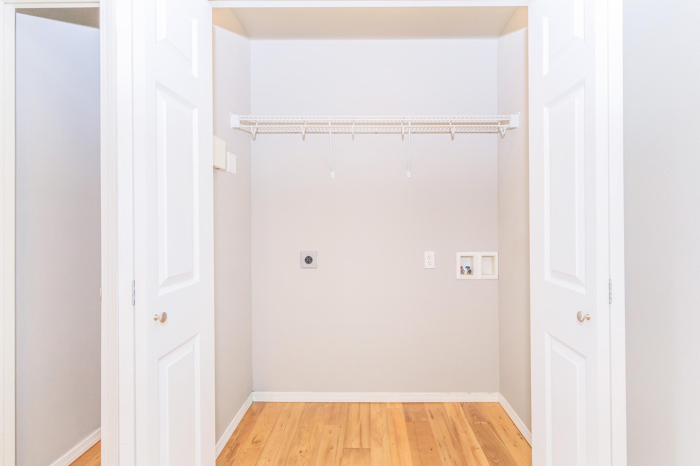 Empty closet with light gray walls and wooden flooring, featuring two white doors that are open. There is a wire shelf mounted against the back wall, and small utility openings are visible on the right side. The space is uncluttered and well-lit.