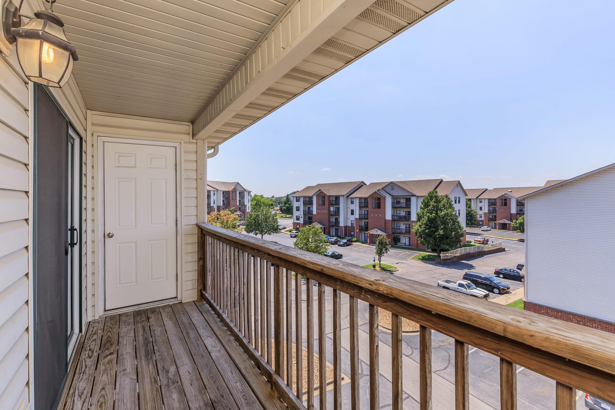 View from a balcony overlooking a parking lot and several apartment buildings. The wooden railing frames the scene, and a door is visible on the left side. The sky is clear with a hint of blue, creating a bright and inviting atmosphere.