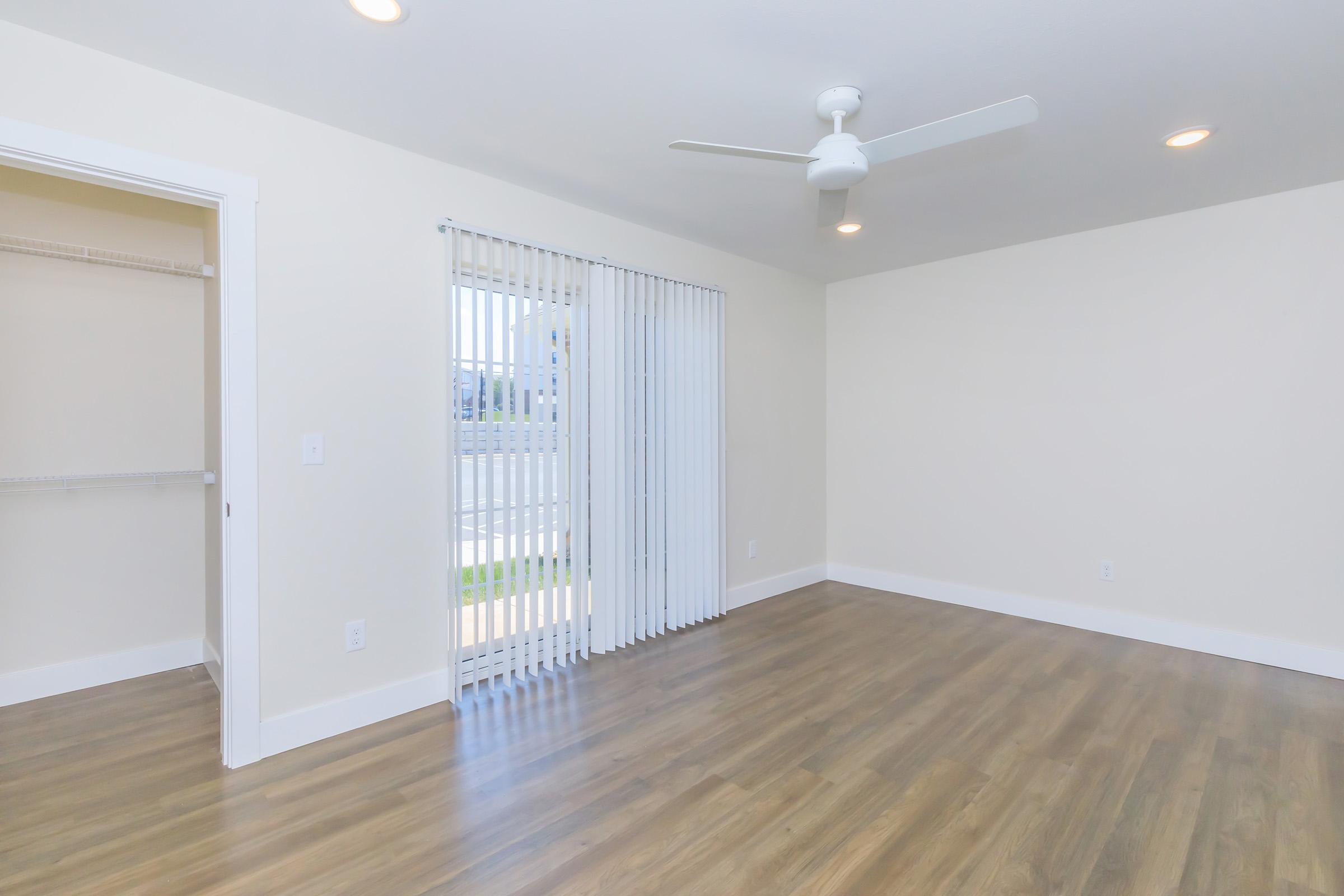 A spacious, empty room with light-colored walls, a ceiling fan, and large sliding glass doors with vertical blinds. There is a small closet with shelves on the left side. The flooring is a light wood laminate, creating a bright and airy atmosphere.