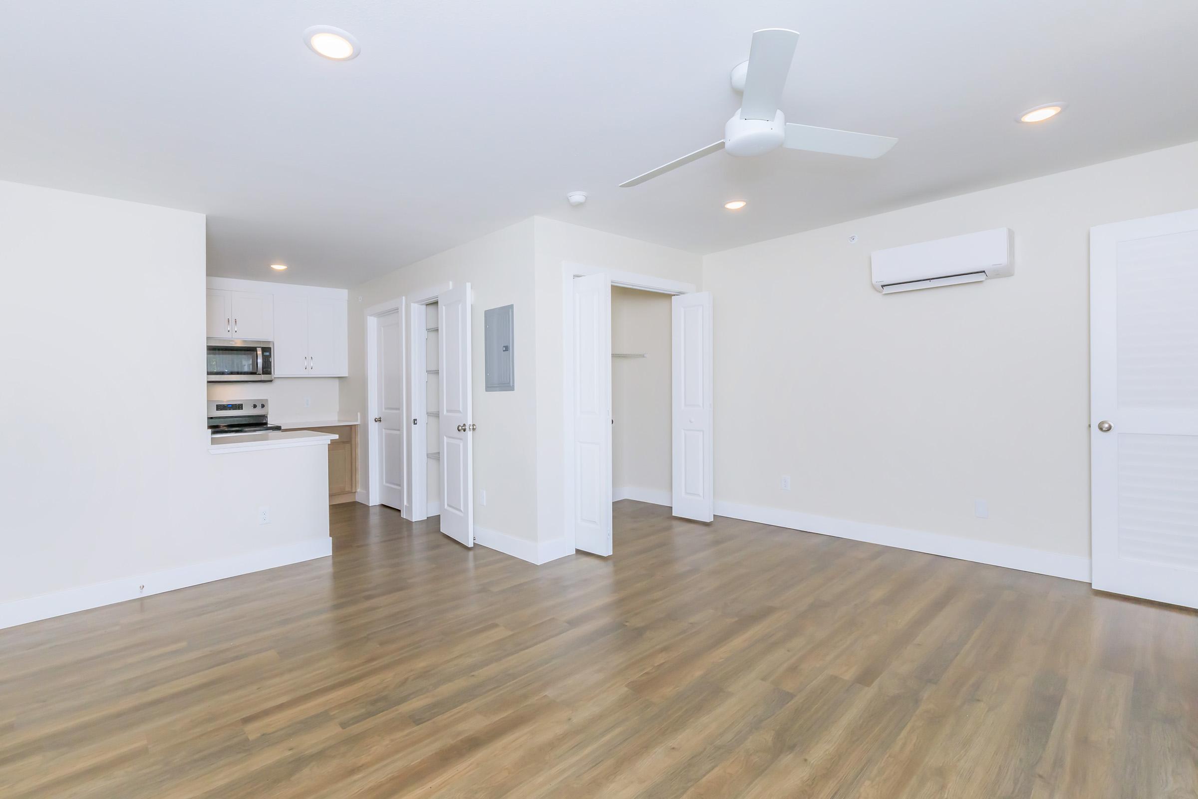 A bright, open living space featuring light-colored walls, laminate flooring, and a ceiling fan. To the left, there's a kitchen area with cabinets and a microwave. Two doorways lead to separate rooms, while a wall-mounted air conditioning unit is visible. The overall atmosphere is clean and modern.