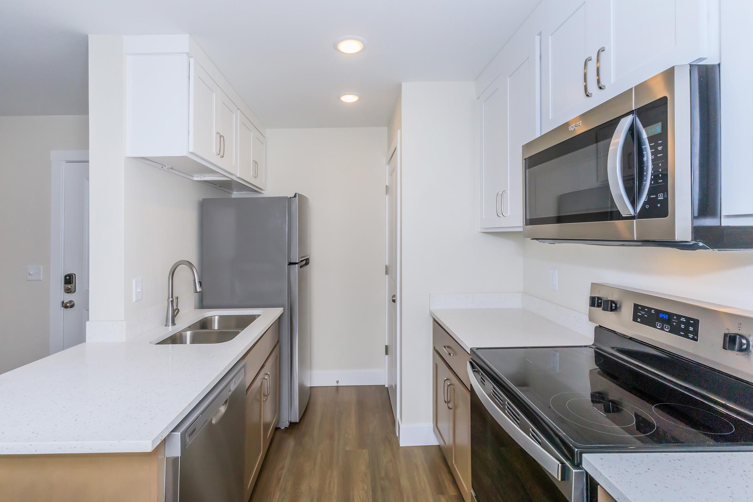 Modern kitchen featuring stainless steel appliances including a refrigerator, microwave, and oven. The cabinetry is white with a sleek design, and there's a double sink under overhead cabinetry. The countertop is a light-colored stone, and the flooring is a warm wood laminate. Soft lighting illuminates the space.