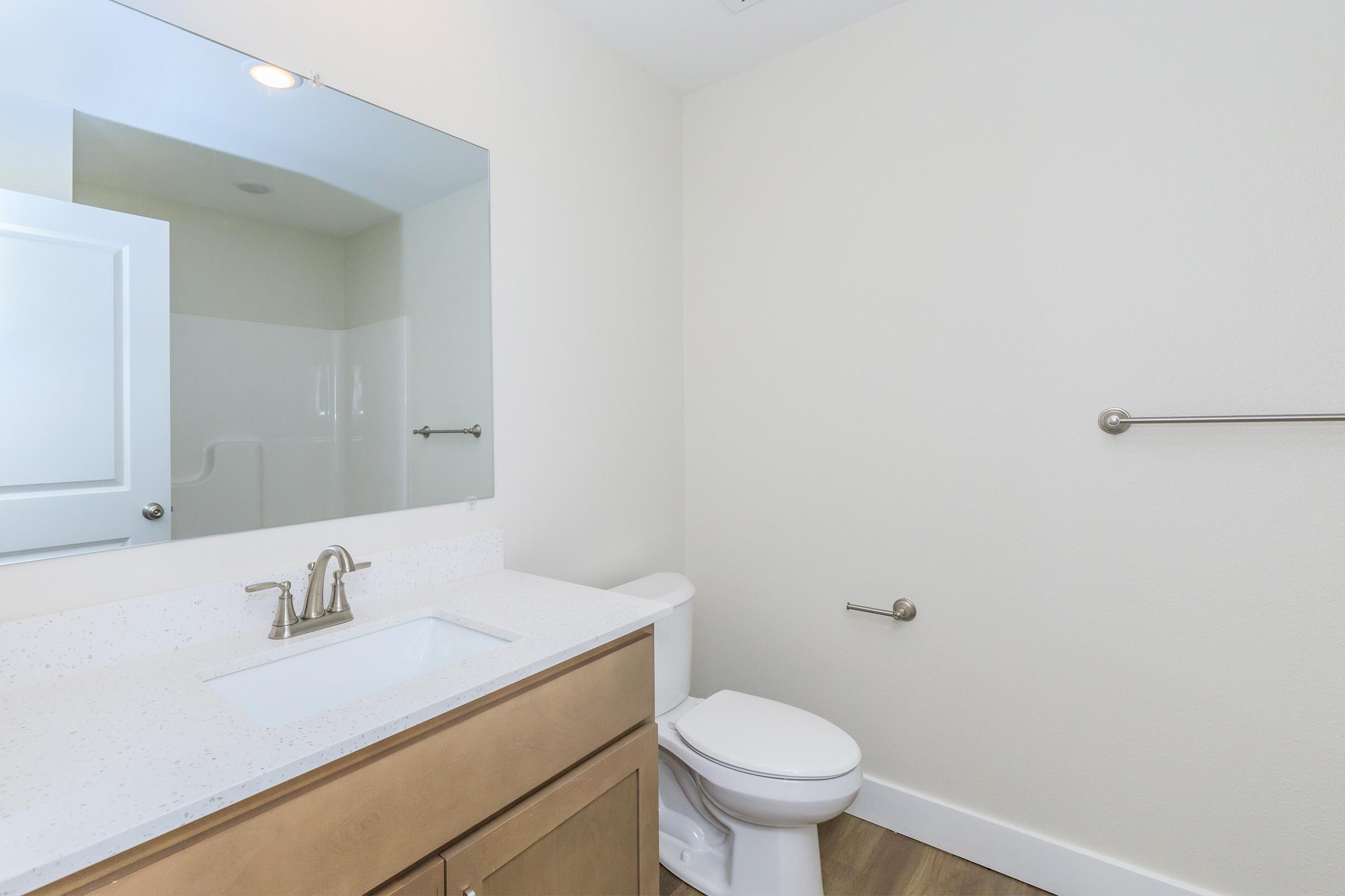 A well-lit bathroom featuring a white countertop sink with a modern faucet, a large mirror above the sink, a toilet, and a light-colored wall. The floor is made of wood-like material, and there is a grab bar mounted on the wall. The space appears clean and contemporary.