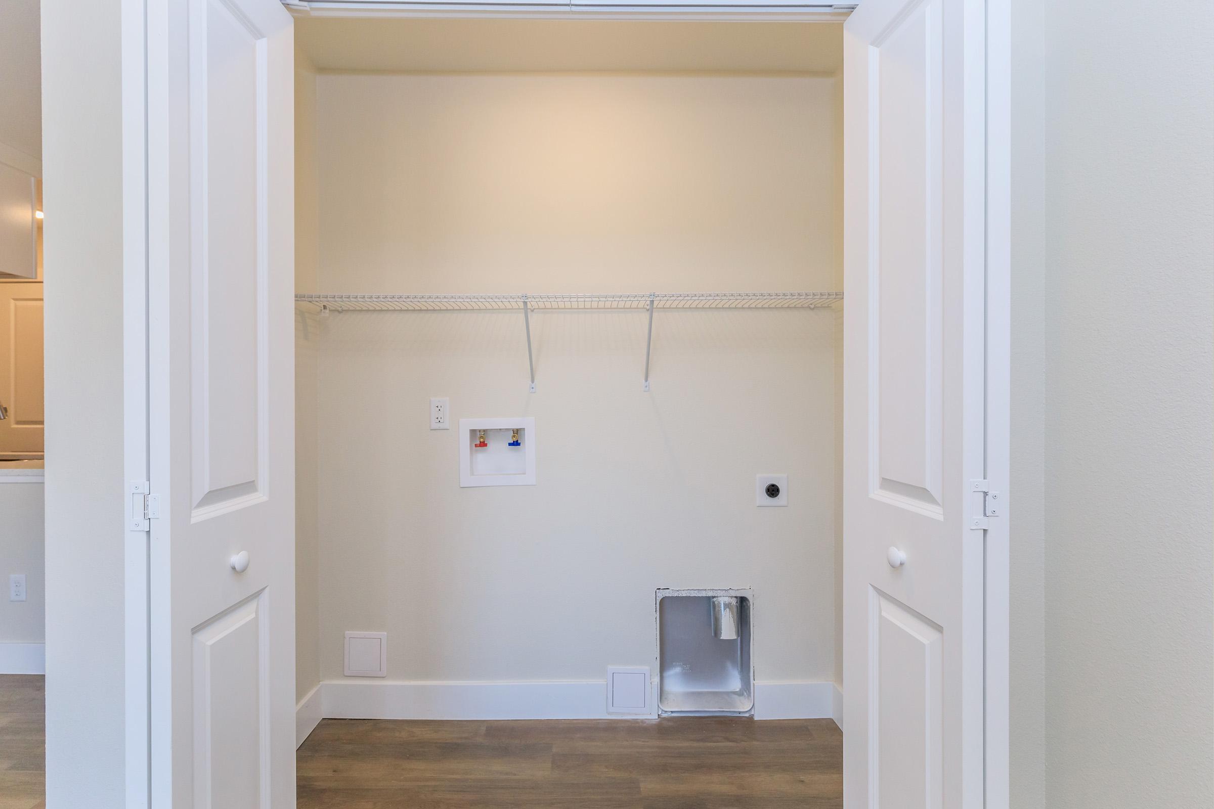 An empty closet with two white doors open wide, revealing a light-colored interior. A wire shelf runs across the top, and there is a small, enclosed space on the floor with a hole, likely for a washer or dryer hookup. The walls are a soft, neutral color, and there is ample natural light in the room.