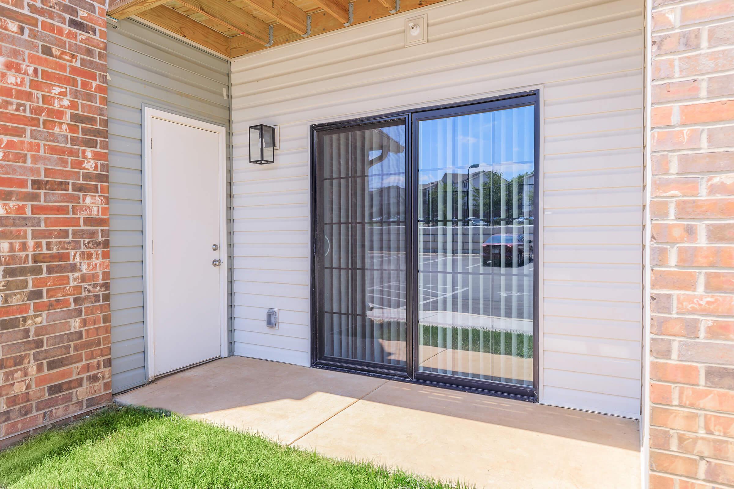 Exterior view of a residential entrance featuring a sliding glass door with black grid pattern, adjacent to a white door. The wall is clad in light-colored siding, while the area around the doors is paved with concrete. Green grass is visible in front of the entrance, and parking is seen in the background.