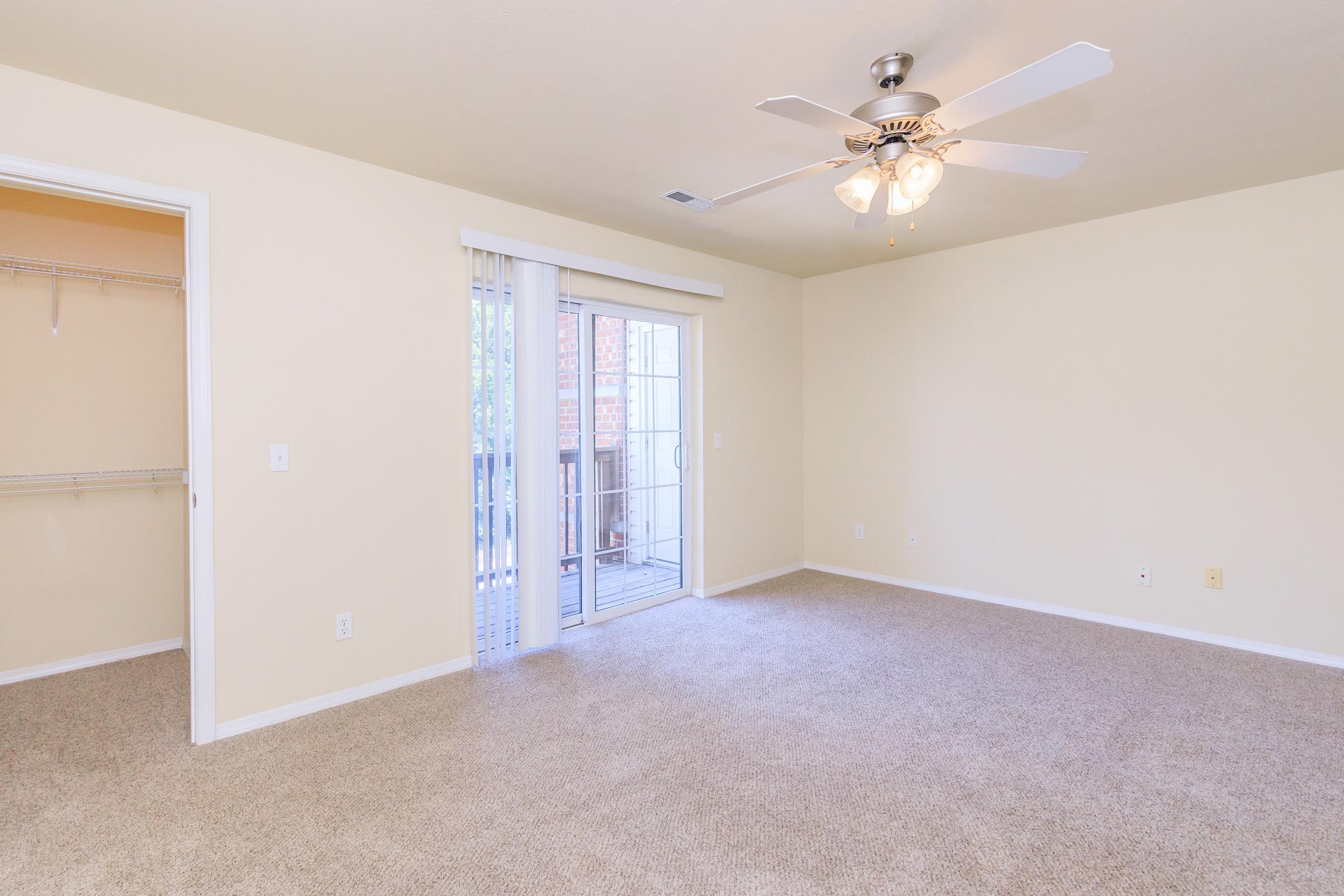 Spacious living room with beige walls and carpet, featuring a ceiling fan and sliding glass doors leading to a balcony. An open closet is visible on the left, creating a bright and airy atmosphere. Overall, the room is well-lit and inviting.
