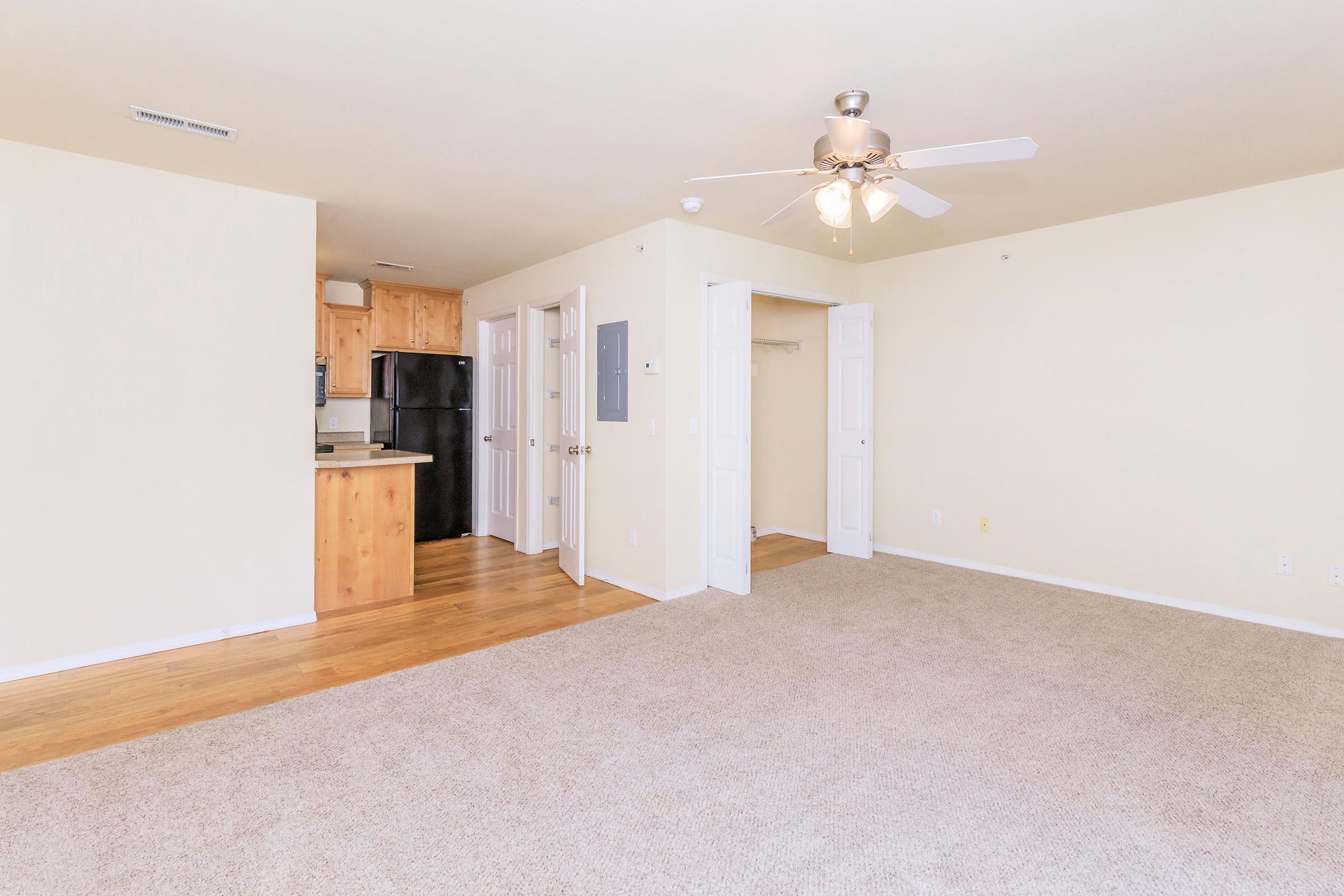 A spacious, unfurnished apartment interior featuring beige walls, a ceiling fan, and a large carpeted area. An open kitchen with wooden cabinetry is visible in the background, alongside a black refrigerator. A closet with double doors is on the right, and the overall ambiance is bright and airy.
