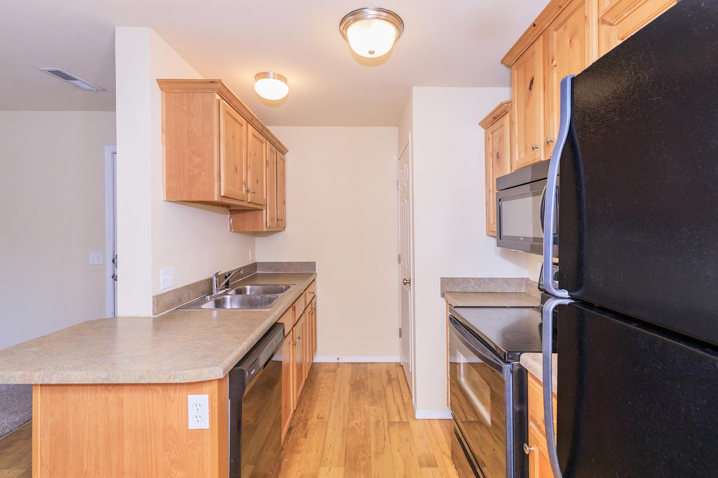 Modern kitchen featuring wooden cabinets, a double sink, and stainless steel appliances including a refrigerator, microwave, and oven. The countertops are made of a neutral stone, and the flooring is a light wood. The lighting includes two ceiling fixtures, providing a bright and inviting atmosphere.