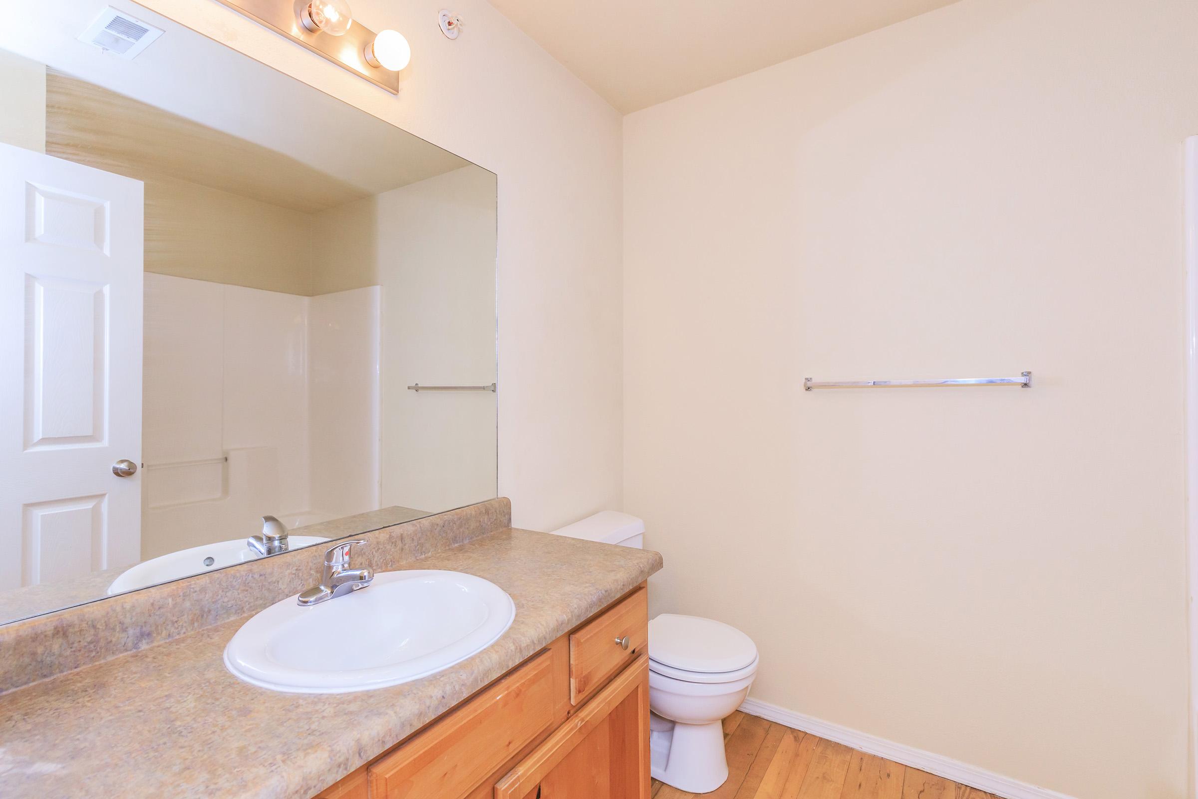 A clean bathroom featuring a wooden vanity with a sink and faucet, a large mirror above, a toilet, and an empty towel rack on the wall. The walls are painted a neutral color, and the flooring is hardwood. Natural light illuminates the space.