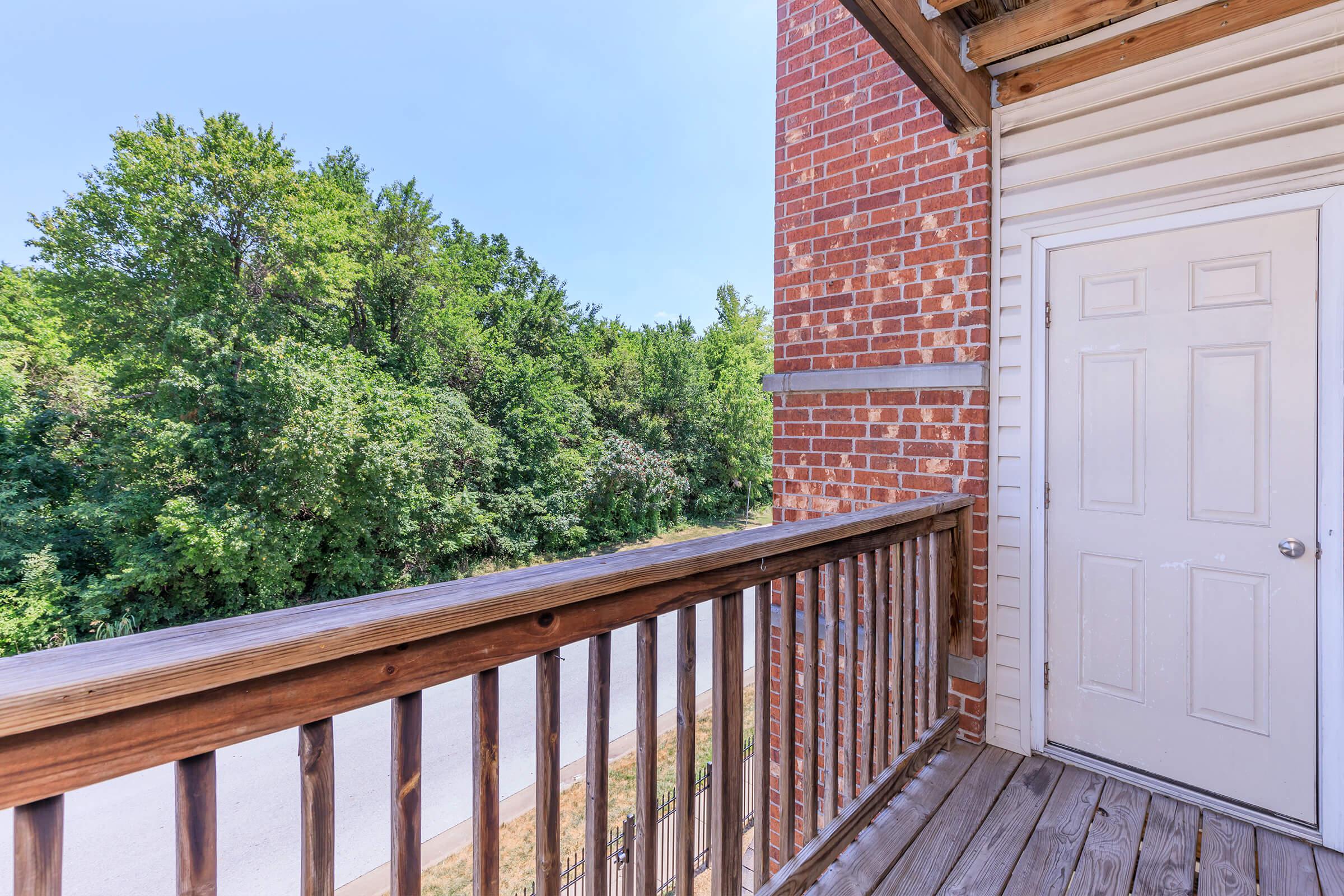 A wooden balcony with a view of lush green trees and a pathway. The brick wall of a building is visible on the right, and a closed white door with a metal handle is located at the back of the balcony. The scene is bright and sunny, creating a peaceful outdoor atmosphere.