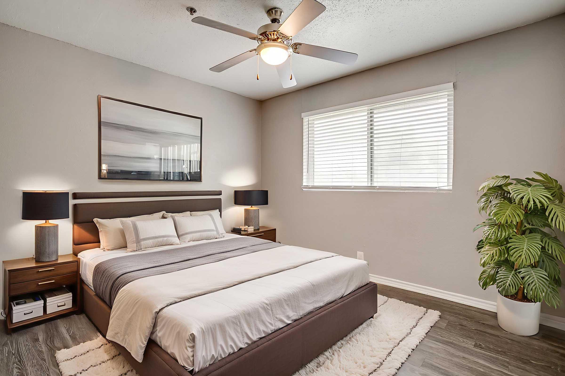 A modern bedroom featuring a neatly made bed with gray and white bedding, two bedside tables with lamps, a large wall art piece above the bed, a window with white blinds letting in natural light, and a potted plant in the corner. The walls are painted in soft neutral tones, adding to the cozy ambiance.