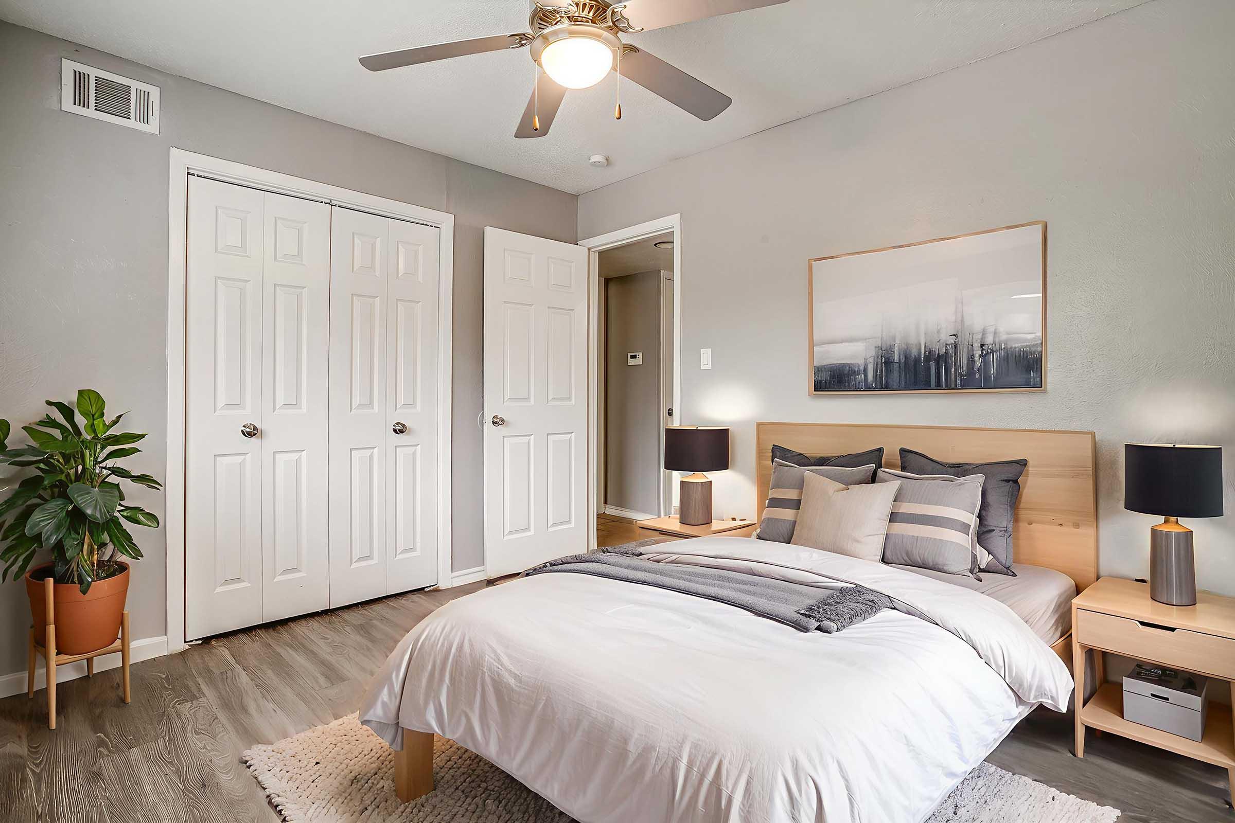 A cozy bedroom featuring a bed with gray and white bedding, two bedside tables with black lamps, and a potted plant in the corner. The walls are painted light gray, and there are two closed doors in the background, one leading to a closet. A ceiling fan adds a modern touch to the room.