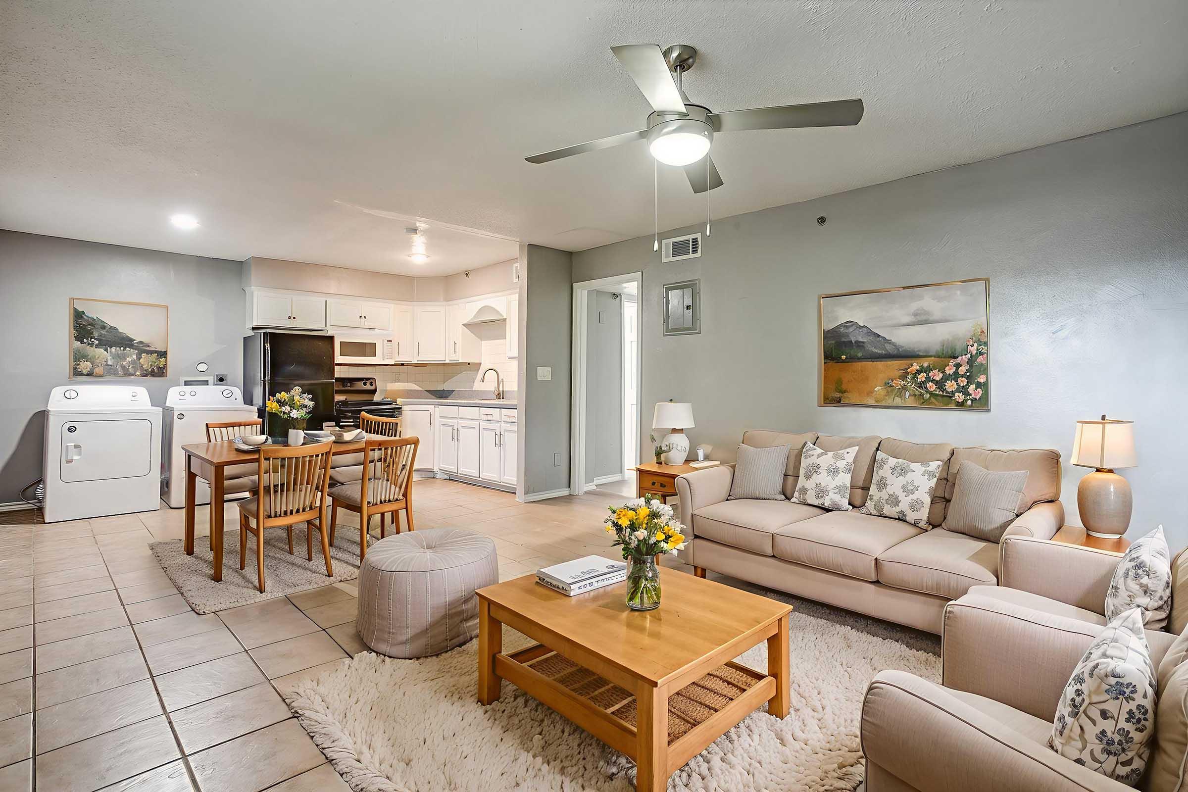 A cozy living room featuring a light-colored couch with decorative pillows, a wooden coffee table, and a pouf. A small dining area with a table and chairs is visible in the background, along with a kitchen equipped with appliances. Laundry machines are also present. The walls are adorned with art, and a ceiling fan is above.