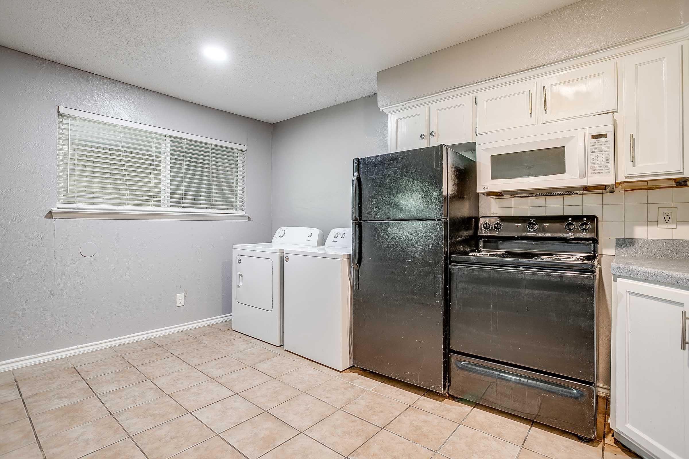 A bright, modern kitchen featuring a black refrigerator, a black oven with a stove, a microwave above, and a white washer and dryer unit. The light-colored cabinets contrast with the gray walls, and there is a window with blinds allowing natural light to enter the room. The floor is tiled in a neutral color.