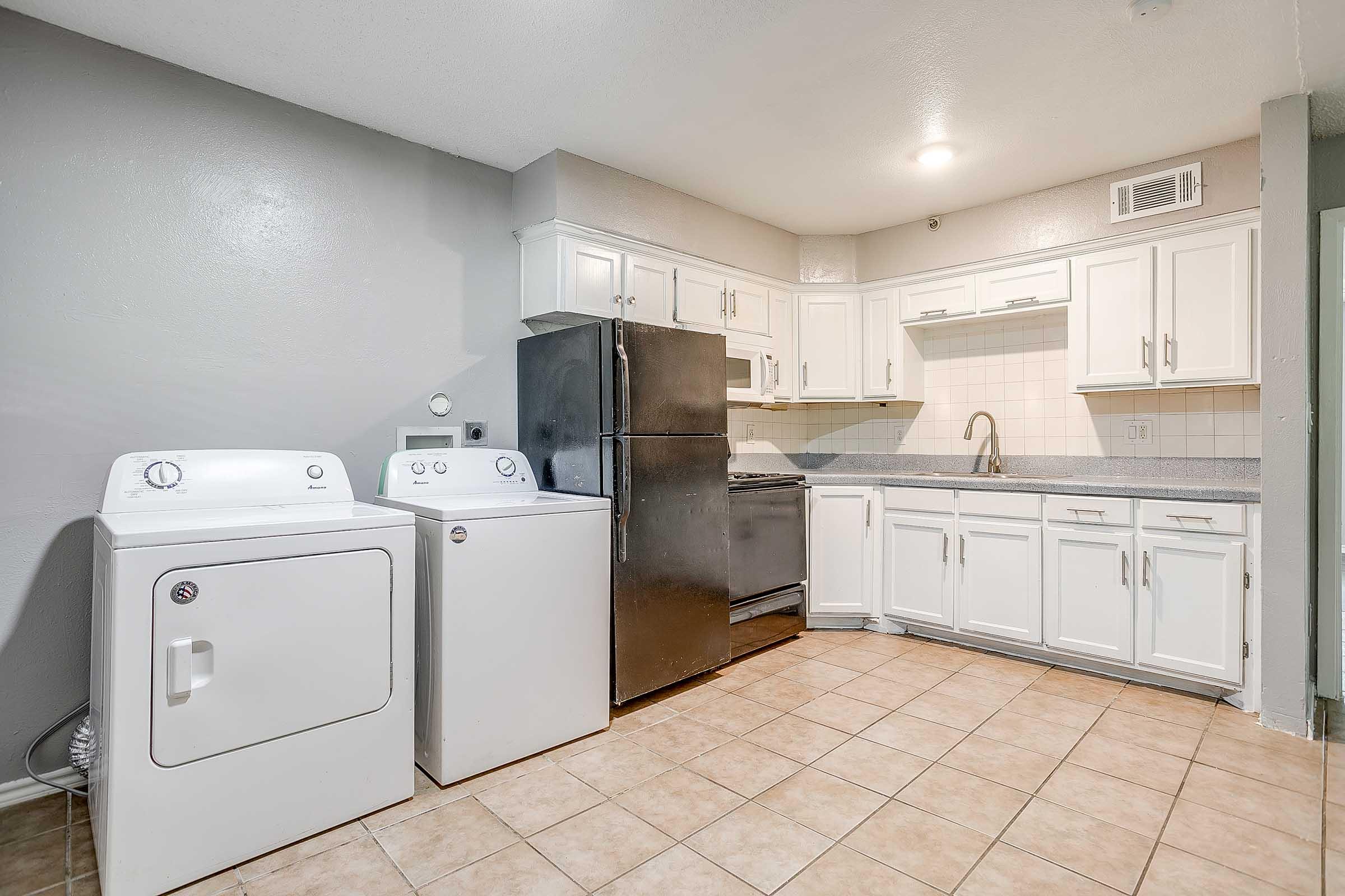 A clean and modern kitchen featuring white cabinetry, a stainless steel sink, a black refrigerator, and a stove. Laundry machines, a washing machine, and dryer, are positioned next to the kitchen area. The flooring is tiled, and the walls are painted in a light gray color.