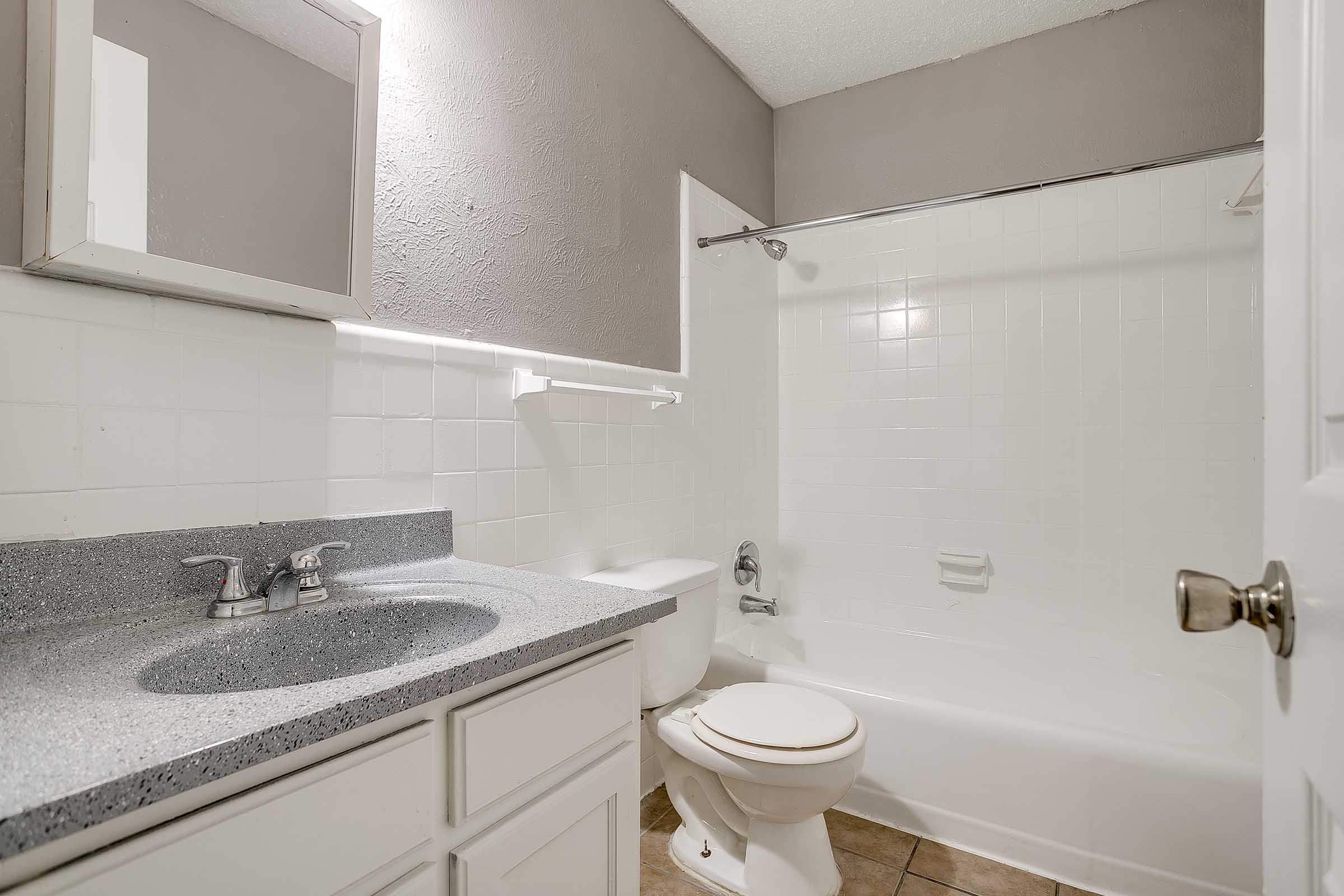 A clean and modern bathroom featuring a white bathtub and shower combination, a toilet, and a sink with a gray countertop. The walls are painted in a light gray color, and there is a small mirror above the sink. Tile flooring completes the space, giving it a fresh and functional appearance.
