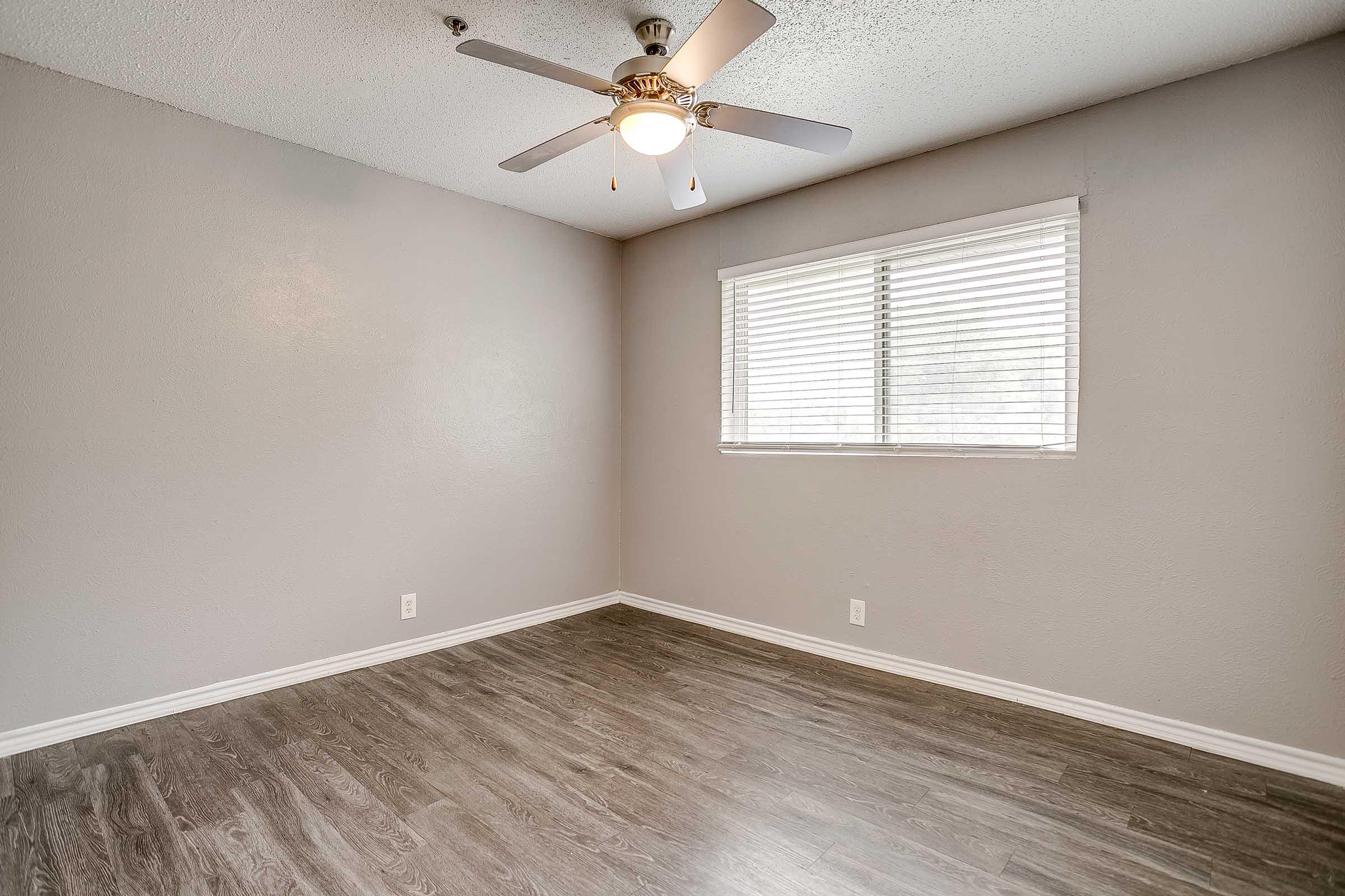 Empty room with light gray walls and a ceiling fan. There is a window with blinds letting in natural light, and the floor is made of dark wood. The room has no furniture or decorations, creating a minimalistic and spacious feel.