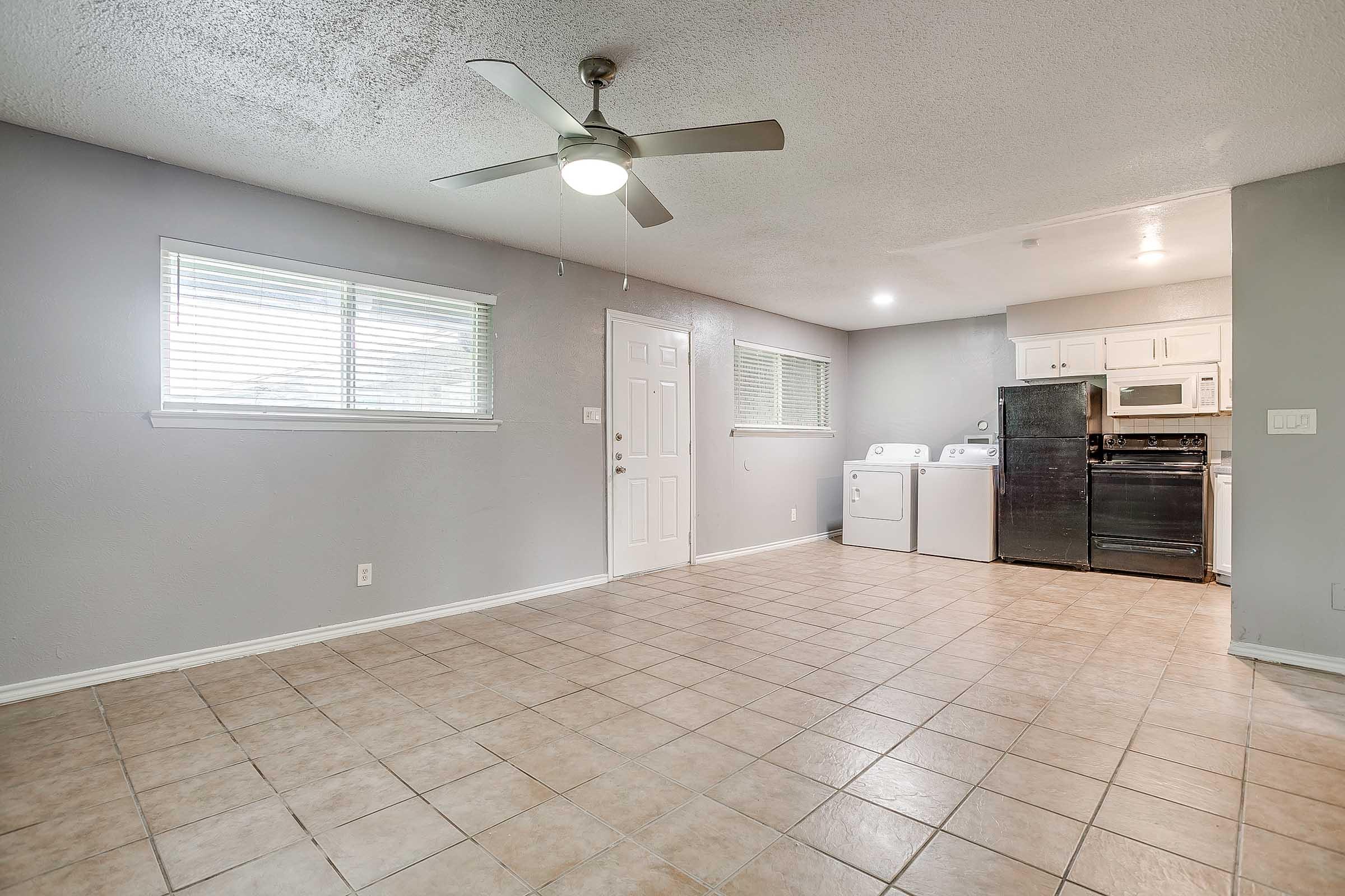 A spacious, well-lit living area with tiled flooring, featuring a ceiling fan. To the right, there is a kitchen area with a refrigerator, stove, and washer/dryer unit. A window allows natural light to enter, and a door leads to another room. The walls are painted in neutral tones, creating a welcoming atmosphere.