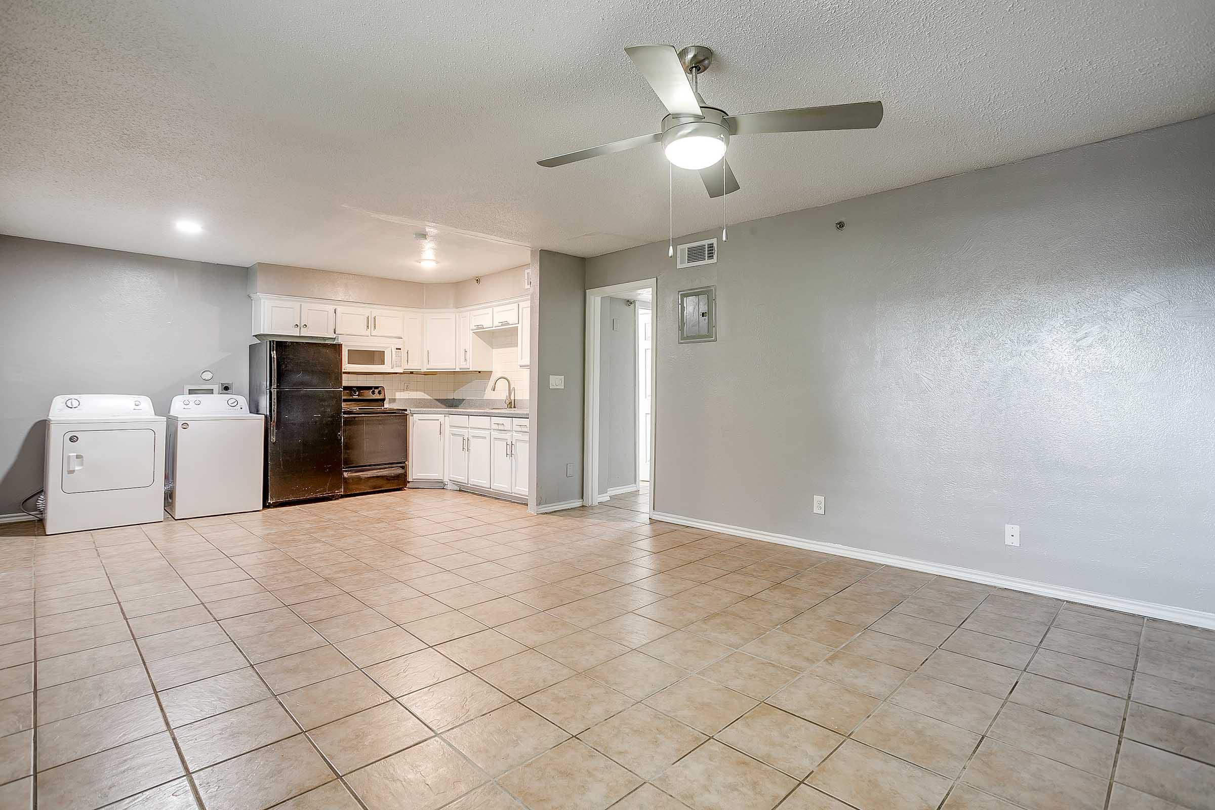 Spacious living area with tiled flooring, featuring a ceiling fan. A kitchenette with white cabinets, a refrigerator, and a black stove is visible. Laundry appliances are along one wall. The room is well-lit with a neutral color scheme and a doorway leading to another area in the background.