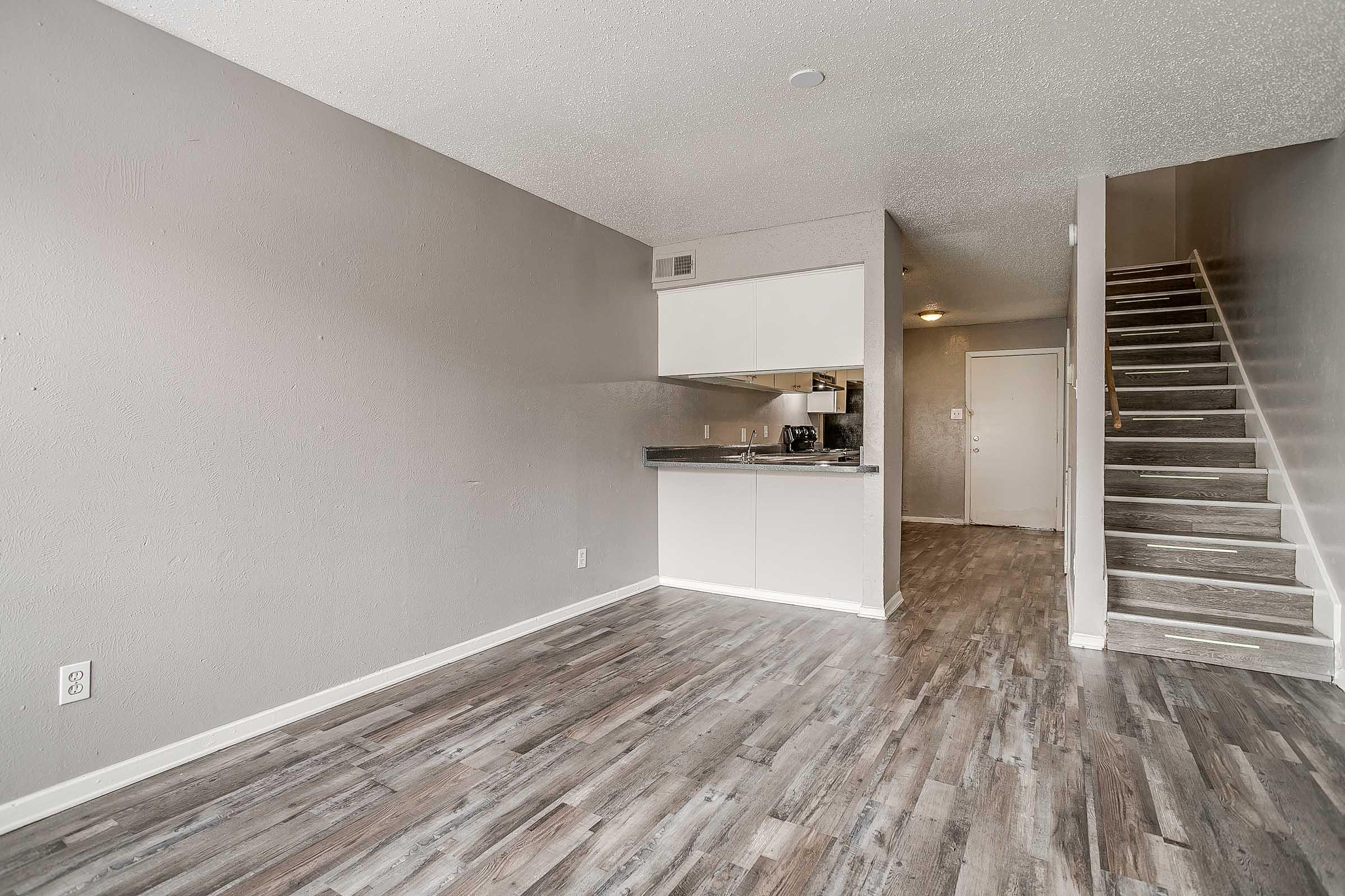 Interior view of a modern apartment featuring an open living space with a kitchen area. The flooring is a light wood laminate, and the walls are painted in a neutral gray tone. A staircase is visible in the background leading to the upper level, while a door can be seen at the far end, suggesting access to additional rooms.