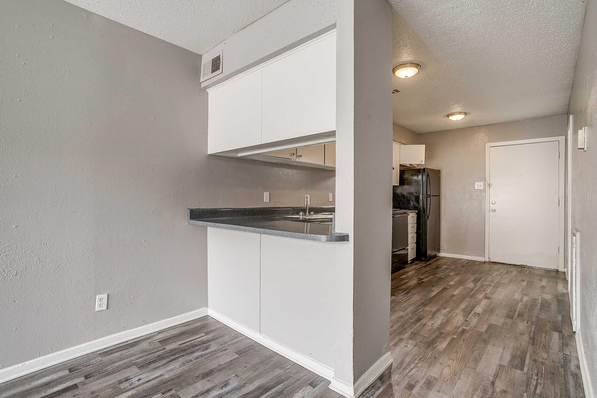 Interior view of a modern kitchen area with gray walls and laminate flooring. The space features a counter with white cabinets above, an entrance door to the right, and an open layout leading to another area. Light fixtures are installed on the ceiling, contributing to a bright atmosphere.