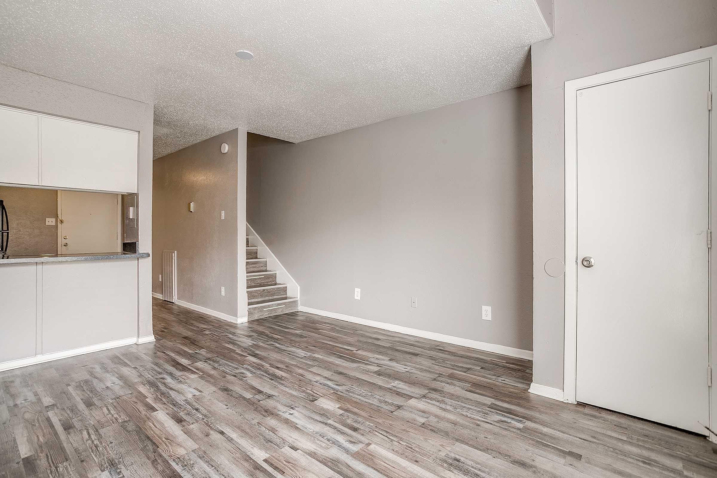 Interior view of a living space featuring gray walls and wood-like flooring. The room includes a staircase on the right, a doorway to the right, and an open kitchen area visible in the background. The space is bright and inviting, with modern finishes and minimal furniture.