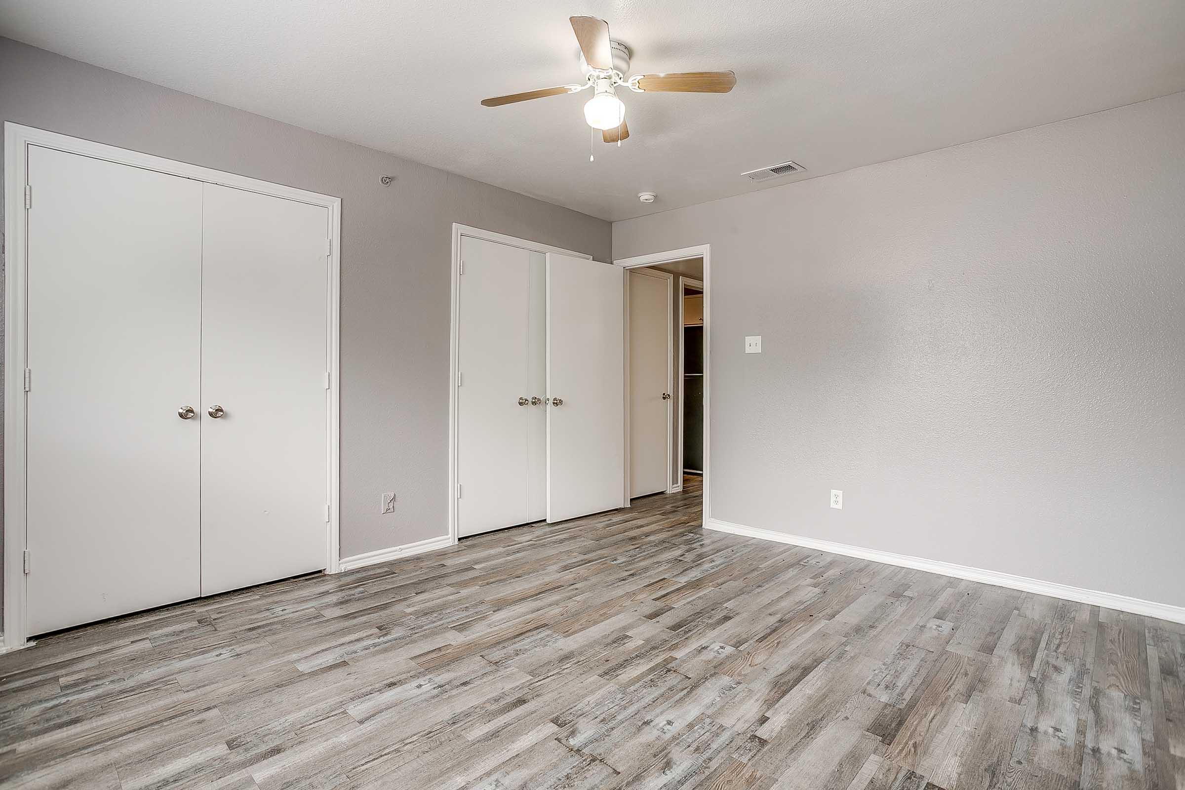 A modest, empty room featuring light gray walls and a ceiling fan. Two white double doors on the left lead to storage or closets, while a doorway on the right provides access to another room. The floor is covered with textured, light wood-like laminate. Natural light brightens the space.