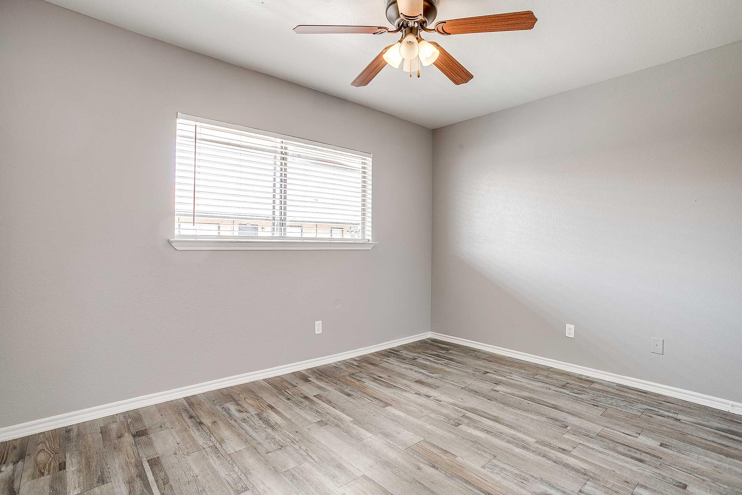 Empty interior of a room featuring light gray walls, a ceiling fan with wooden blades, and a window with blinds. The floor has a wood-like finish, and there are no furnishings present, giving the space a clean and open appearance.