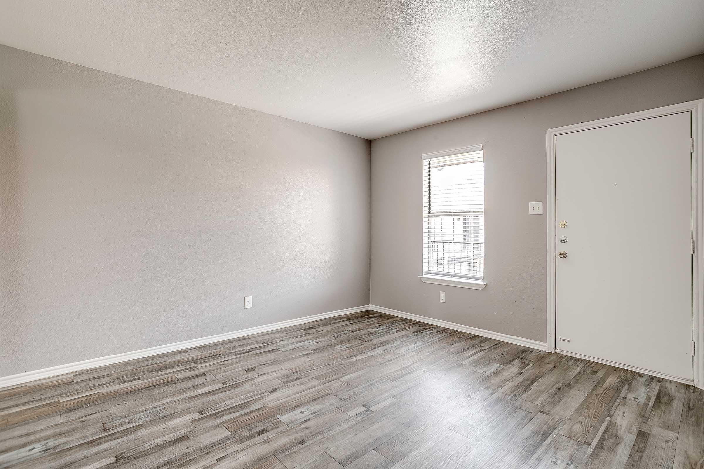 A vacant room with light gray walls, a window with blinds on the left, and a white door on the right. The floor features laminate wood planks, and the space is well-lit with natural light. The overall atmosphere is clean and modern.