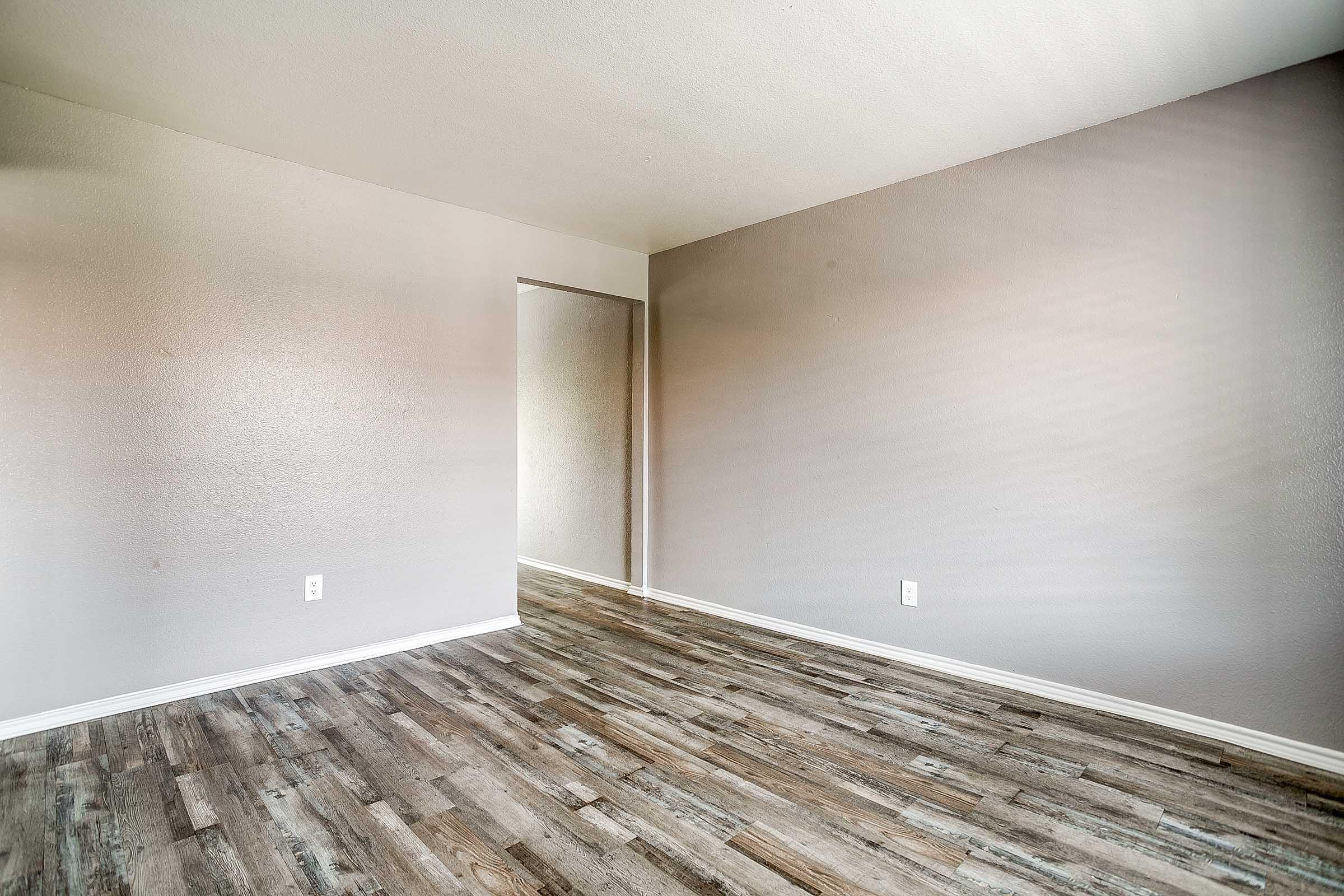 A vacant room with light gray walls and wood-like flooring. The space has a recessed area, possibly for a doorway, and appears bright with natural lighting. No furniture or decorations are present, giving a clean, minimalistic look.