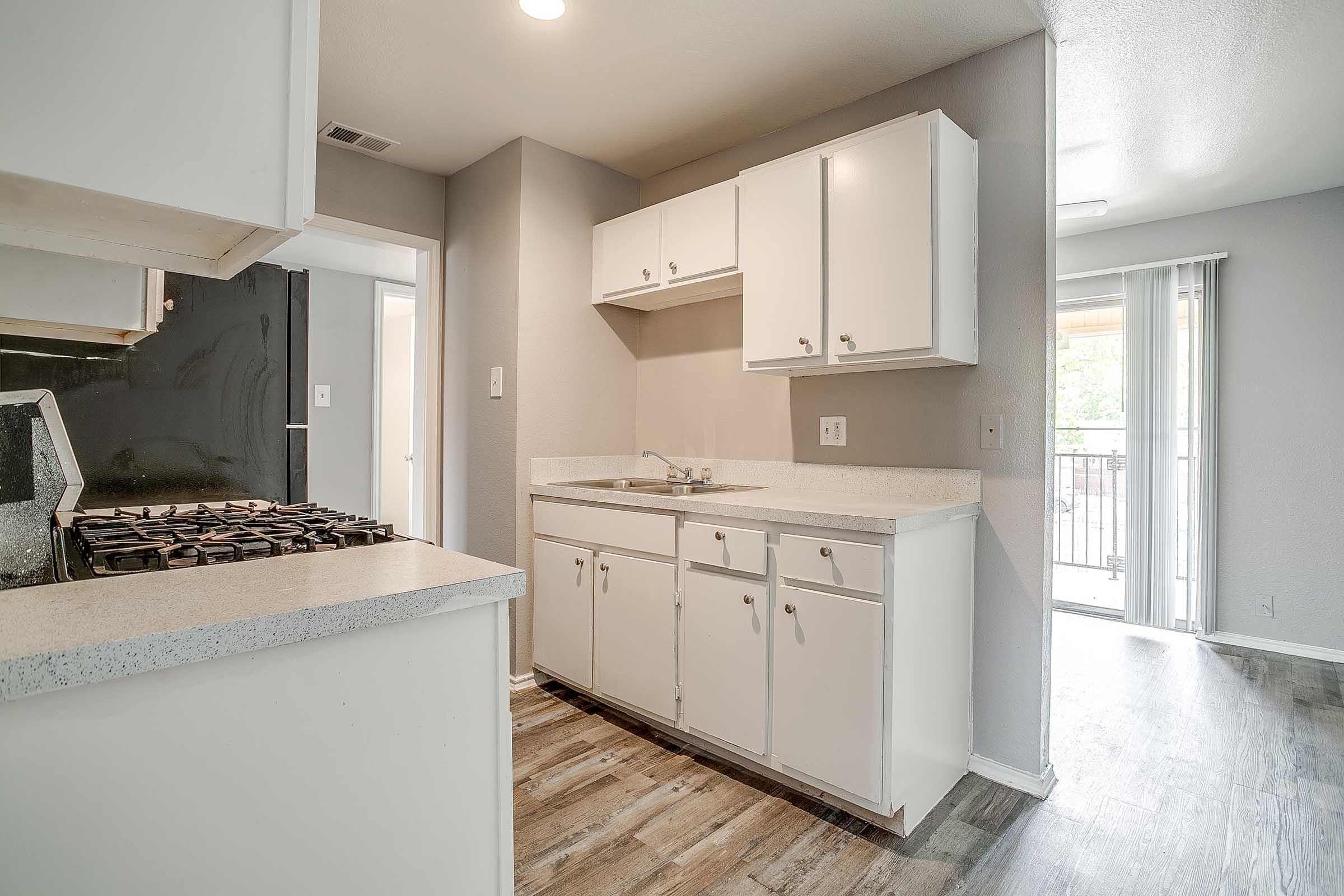A modern kitchen featuring white cabinets, a granite countertop, and a gas stove. The flooring is a light wood laminate, and there's a door leading to a balcony. The walls are painted in a neutral color, and the space is well-lit, creating a bright and inviting atmosphere.