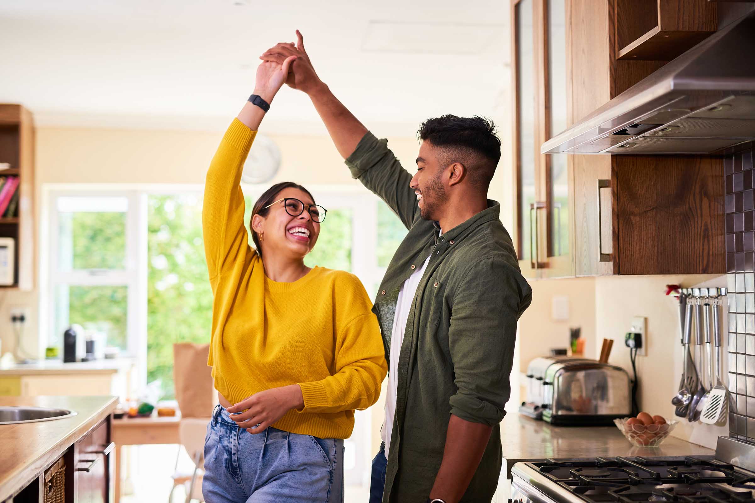 A joyful couple dancing together in a bright kitchen. The woman, wearing a yellow sweater and glasses, smiles broadly as the man, dressed in a green shirt, lifts her hand playfully. Sunlight fills the space, enhancing the warm, cheerful atmosphere.