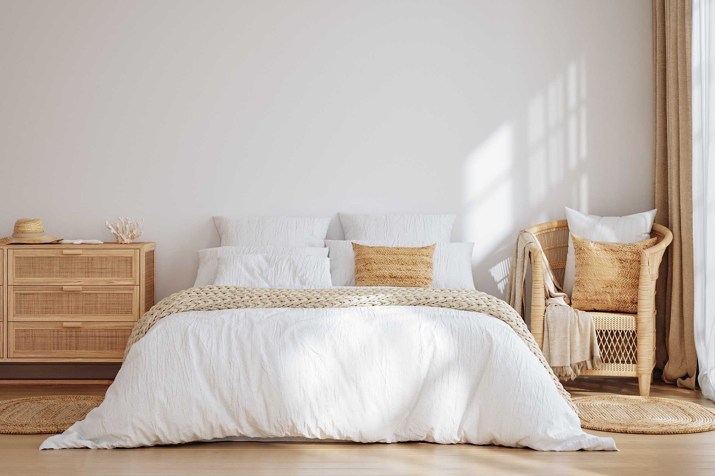 A cozy bedroom featuring a large white bed with textured bedding and decorative pillows, a light wood nightstand, and a woven chair next to a window. Natural light filters through sheer curtains, creating a warm and inviting atmosphere. Subtle earth tones enhance the serene decor.