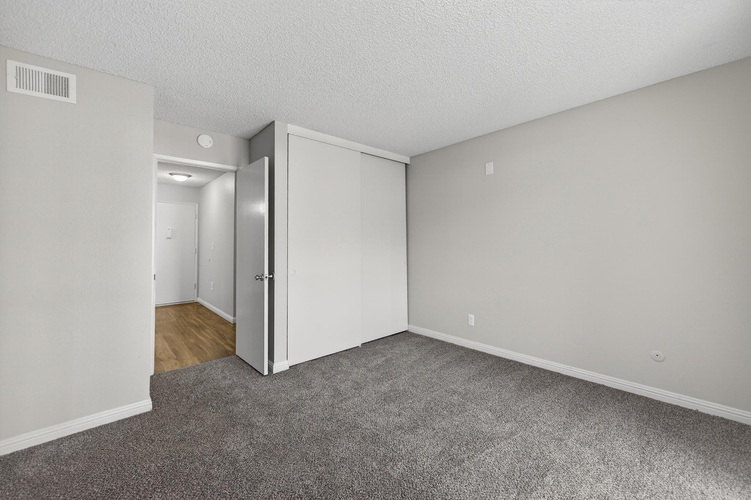 Interior view of a vacant room with light gray walls and plush carpet. A closed white closet door is visible on the left, while an entrance door leads to a hallway on the right. Soft lighting from a ceiling fixture illuminates the space, which has a clean and modern look.