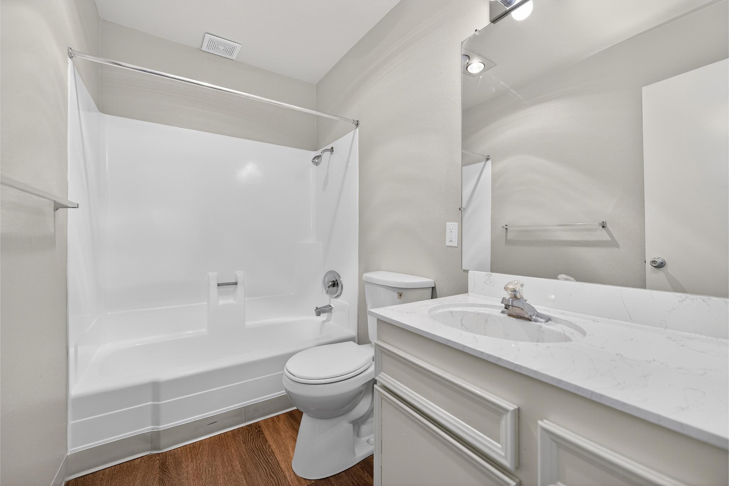 A clean and modern bathroom featuring a white bathtub and shower combination, a toilet, and a sink with a marble countertop. The walls are painted in a light color, and there is a mirror above the sink, with minimalistic lighting. The floor is finished with wooden laminate.