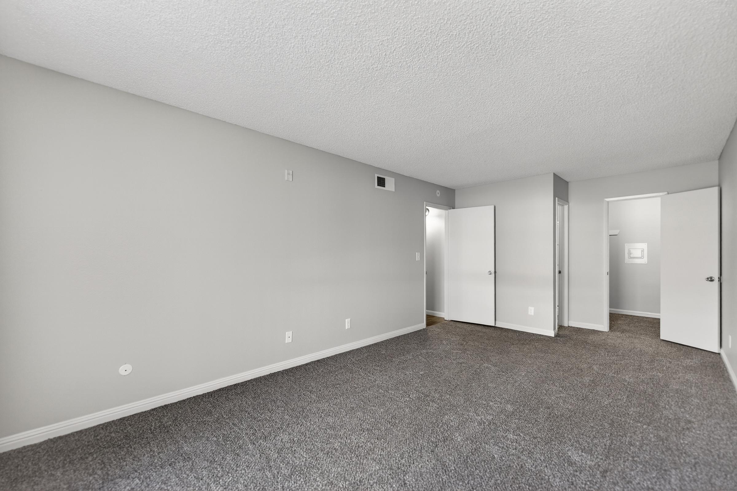 A spacious room with gray walls and carpeted flooring. Two doors are visible, leading to adjacent areas, and the natural light brightens the interior. The room is empty, creating a clean, minimalist appearance suitable for various layouts.