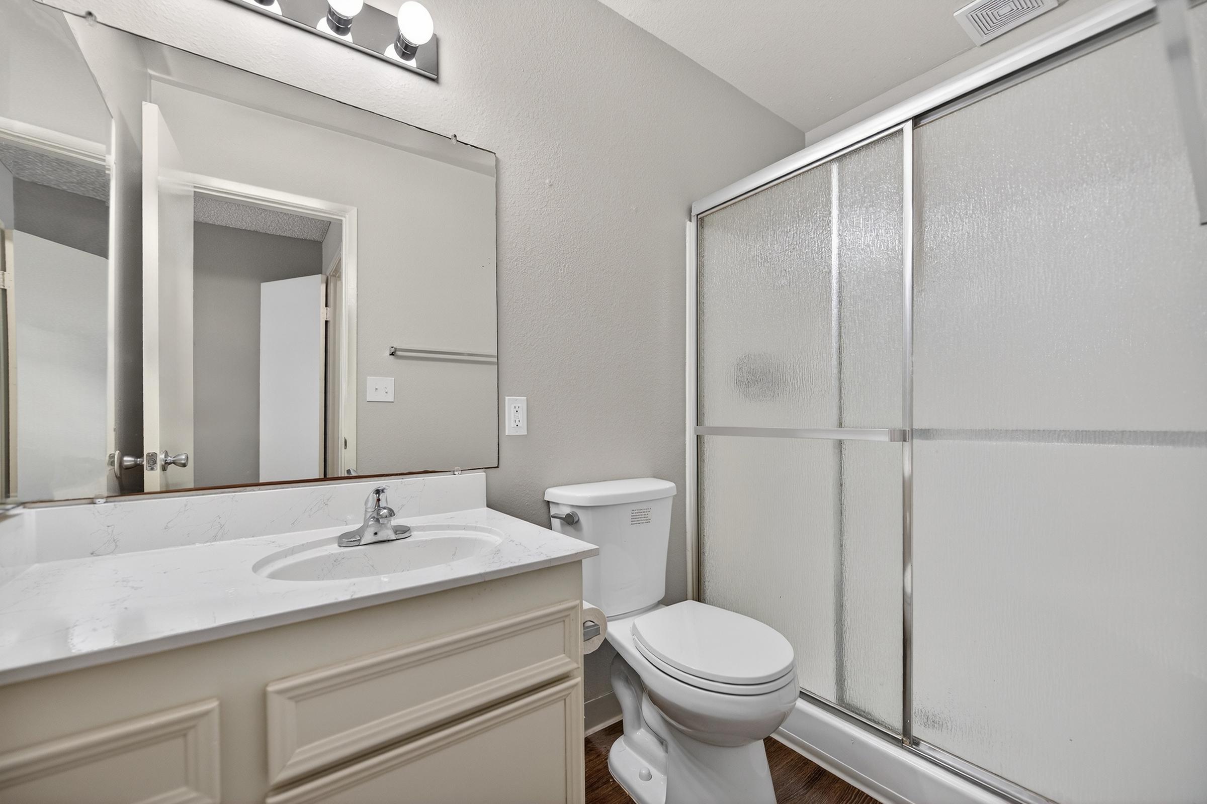 Modern bathroom featuring a light-colored vanity with a sink and mirror, a white toilet, and a glass-doored shower. The walls are painted in a neutral tone, and there is soft lighting from a wall-mounted fixture above the mirror. The floor has a wooden appearance, adding warmth to the space.