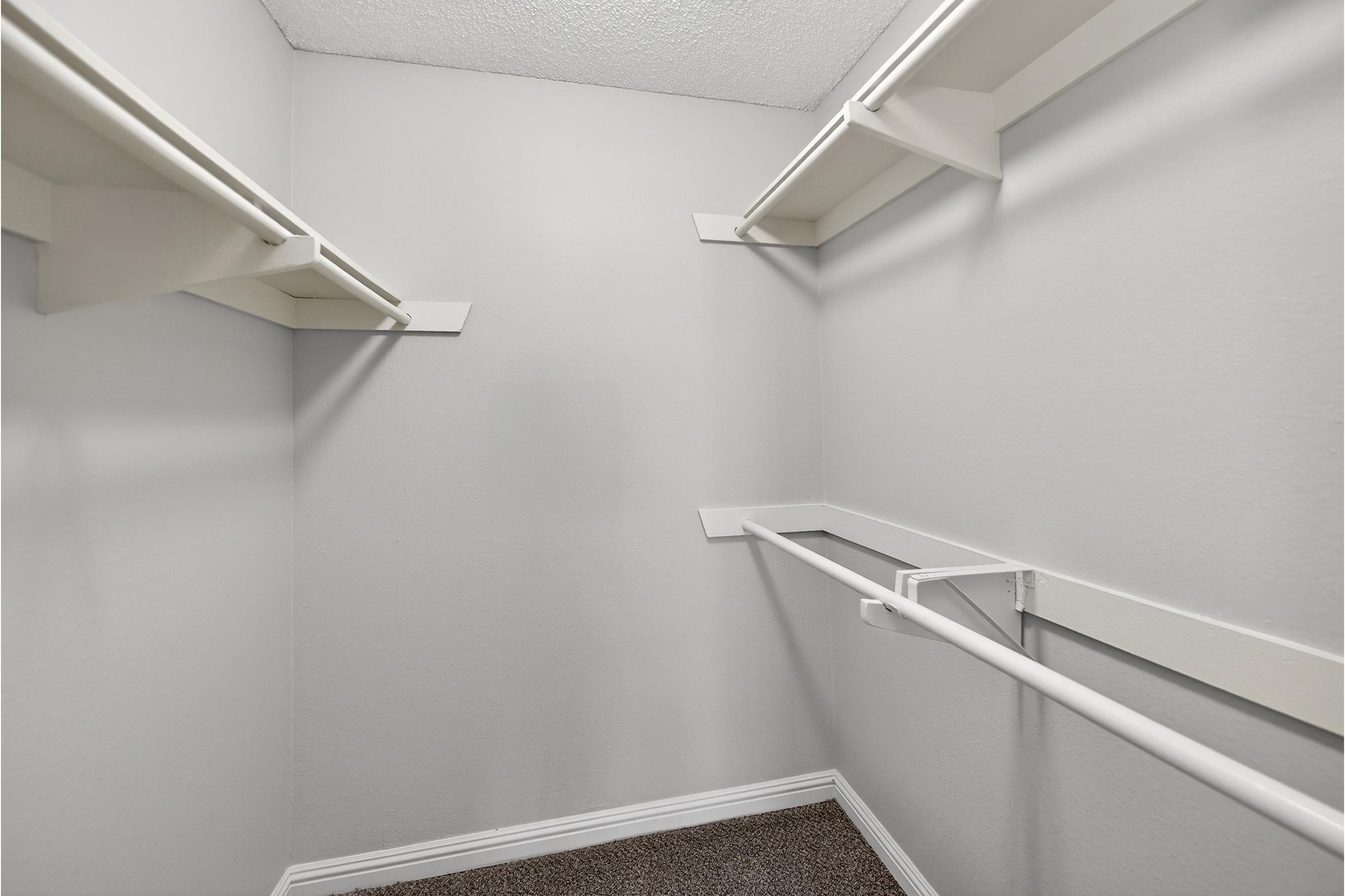A small, empty closet with gray walls, featuring two horizontal shelves on the left side and a hanging rod. The floor is covered with a brown carpet, creating a clean and minimalistic storage space.