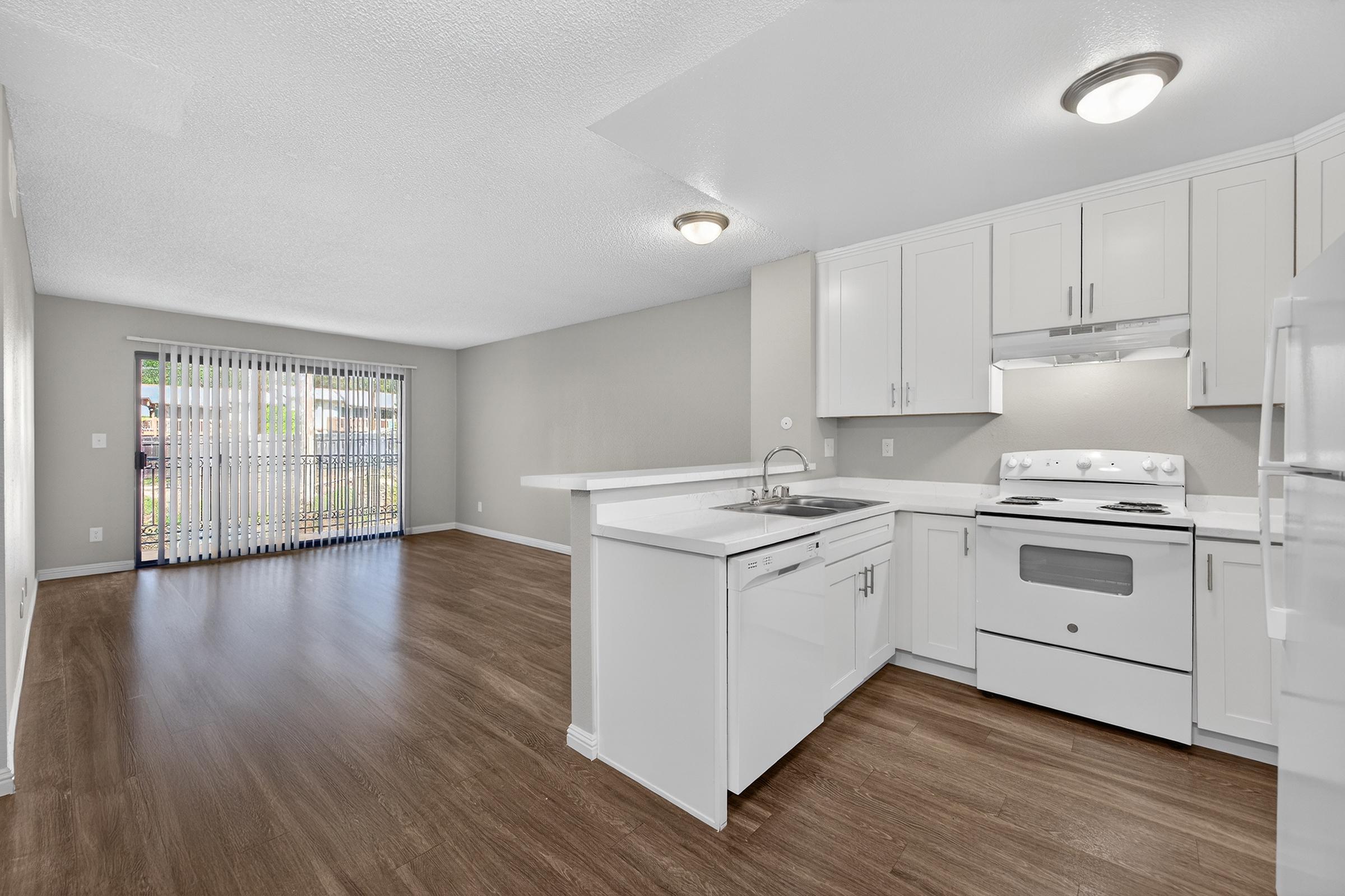 Modern kitchen and living area featuring white cabinetry, a kitchen island with a sink, and stainless steel appliances. The space has wood-style flooring, recessed lighting, and large windows with vertical blinds, allowing natural light to fill the room.