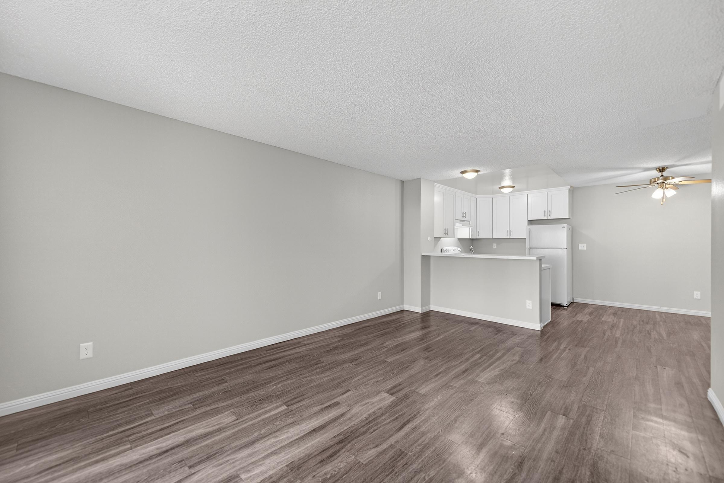 A spacious, empty living room with smooth, dark wooden flooring and light gray walls. In the background, there is a kitchen featuring white cabinets and appliances. A ceiling fan is visible, and the overall lighting is bright, creating a clean and airy atmosphere.