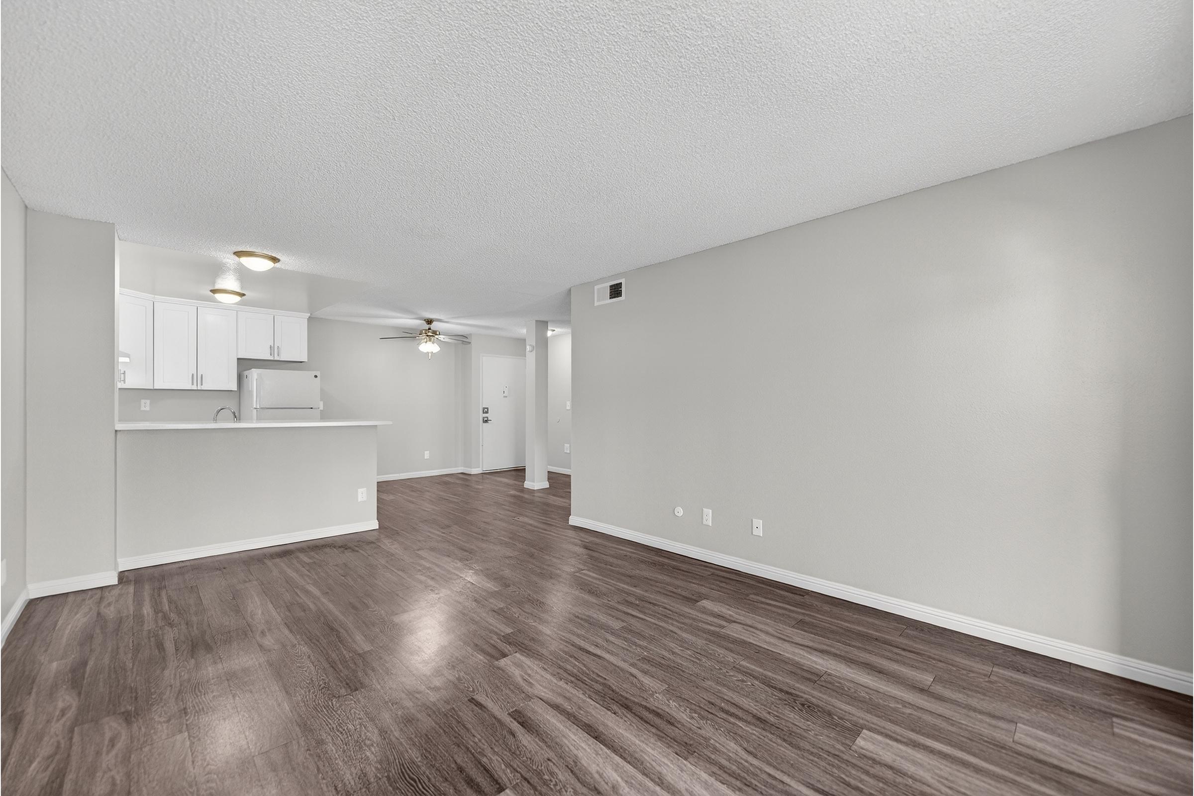 A spacious living area with light-colored walls and dark wood flooring. To the left, there is a kitchen with white cabinets and a counter. The area is well-lit, featuring a ceiling fan and natural lighting from the windows, creating a bright and airy atmosphere.