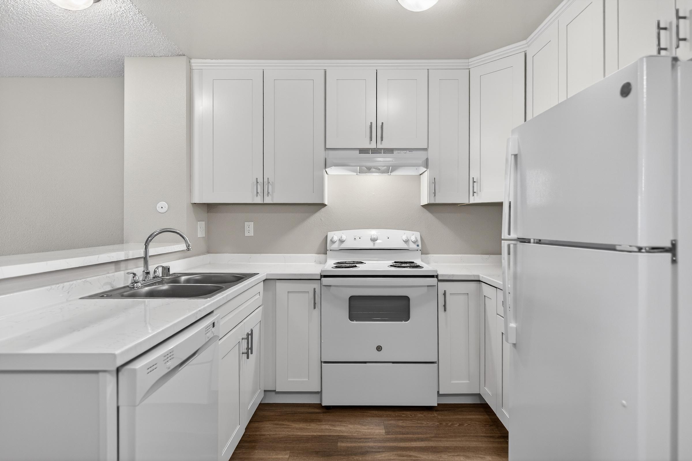 A modern kitchen featuring white cabinets, a double sink, a white refrigerator, and a white stove with an overhead exhaust fan. The countertops are light-colored, and the flooring is wooden. The walls are a neutral color, creating a bright and clean atmosphere.