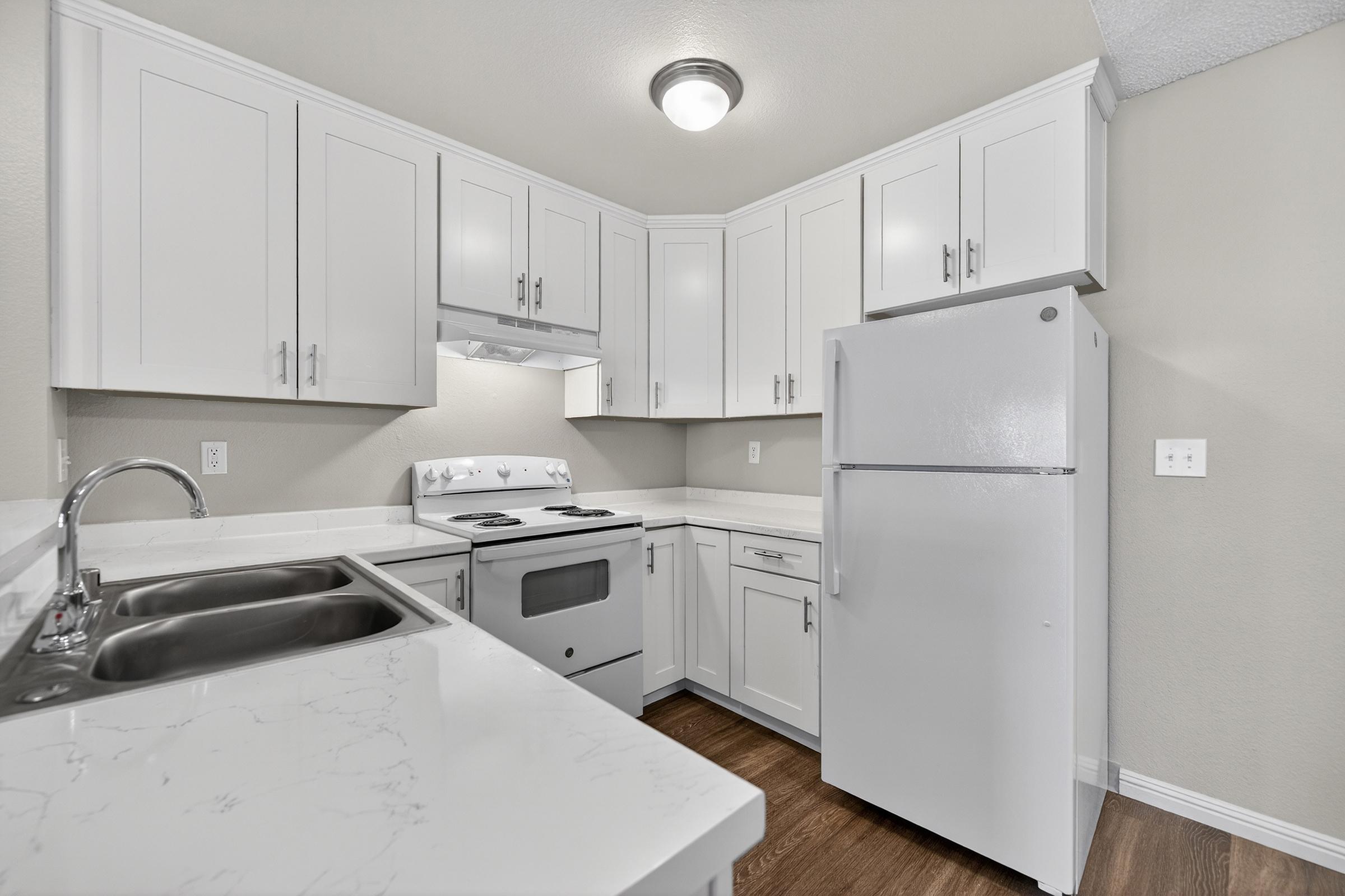 Modern kitchen featuring white cabinets, a white refrigerator, and a white stove. The countertop is light-colored with a marbled pattern. The walls are painted in a soft neutral tone, and there's overhead lighting. The kitchen has a clean, minimalist design with a sink and ample storage space.