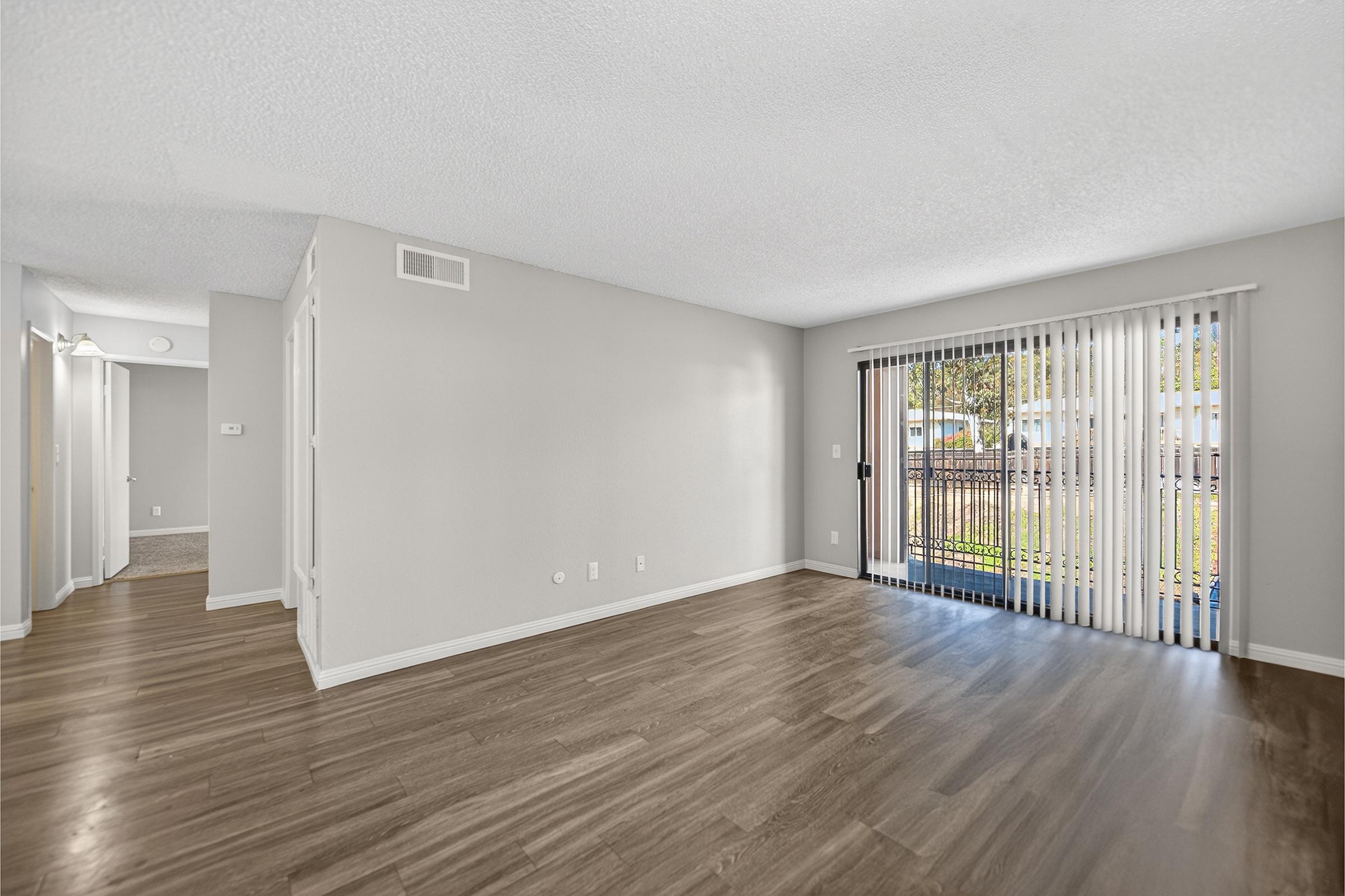 A spacious, empty living room featuring light-colored walls and hardwood flooring. There are large windows with vertical blinds allowing natural light, and a sliding door leading outside. The room is uncluttered and ready for furnishing, with a hallway visible on the left side.