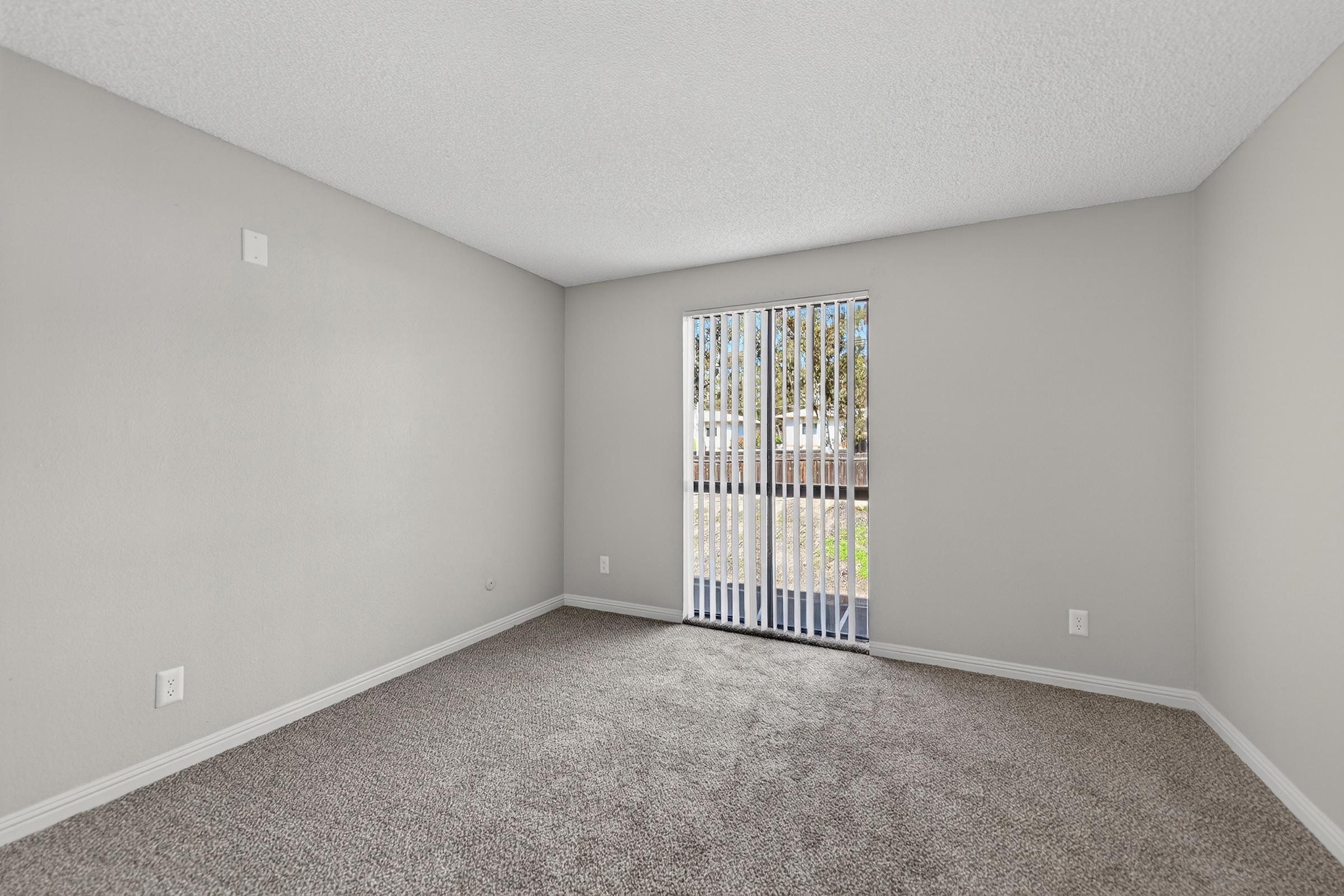 Empty room with light grey walls, carpeted floor, and a window featuring vertical blinds. Natural light is coming through the window, which opens to a balcony with a view of greenery outside.