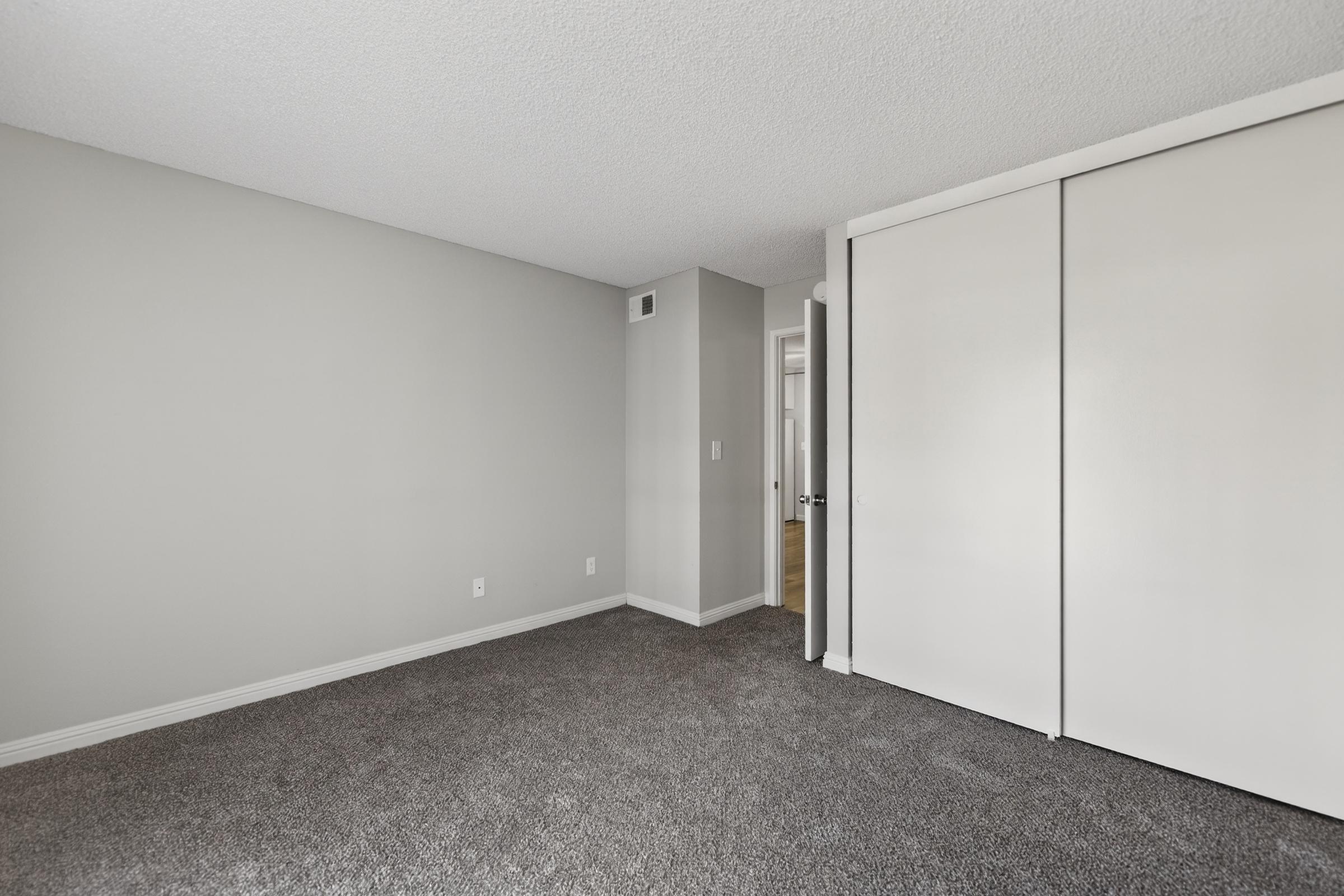 A spacious, empty room with gray walls and a carpeted floor. There is a noticeable closet with sliding white doors on one side and a door leading to another room in the background. Natural light brightens the space, creating a clean and minimalist atmosphere.