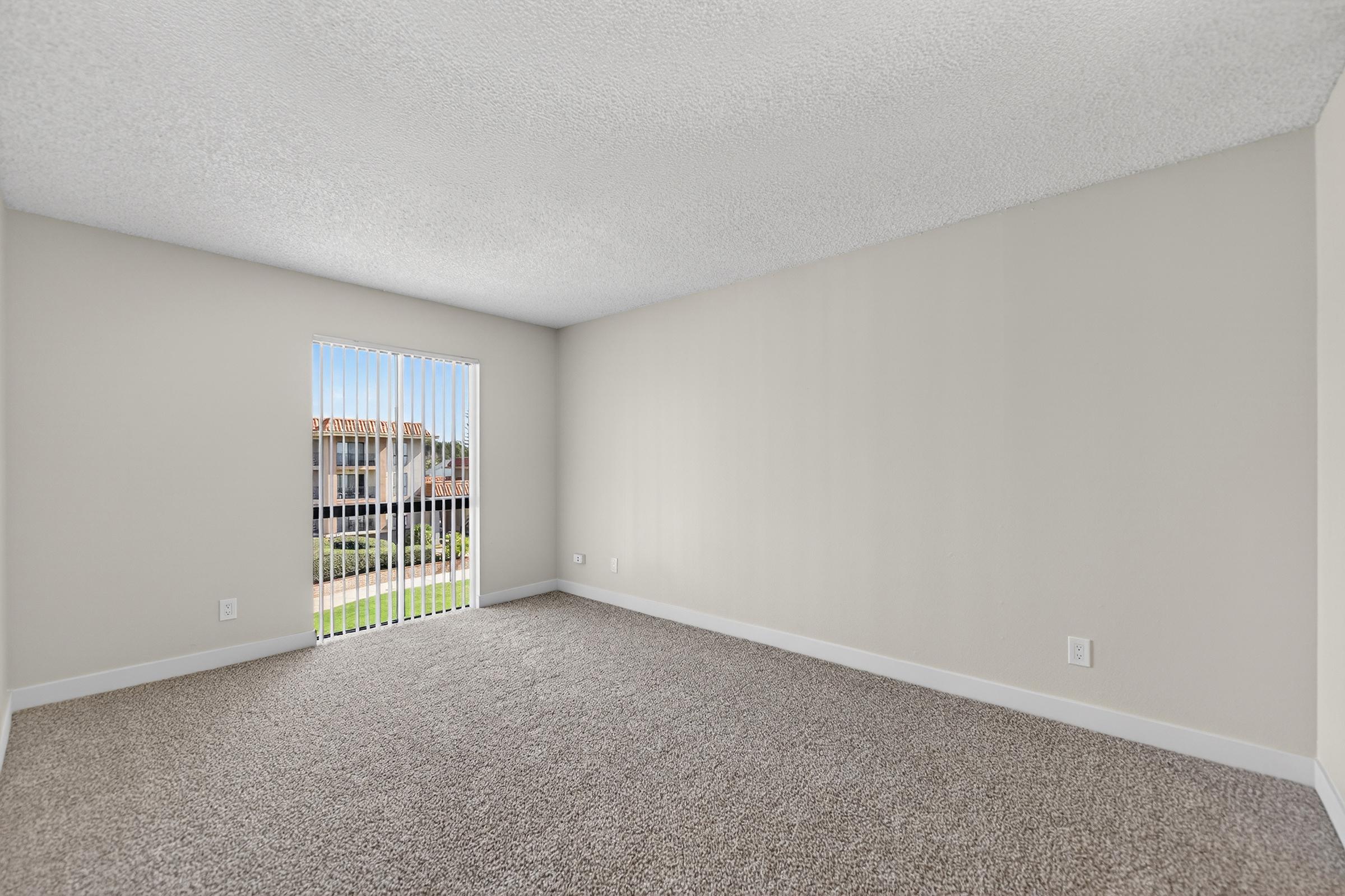 A bright, empty room with beige walls and textured ceiling. The floor is covered in light brown carpet. A large window with vertical blinds lets in natural light and offers a view of the outside, which features buildings or houses. The space appears clean and ready for furniture placement.