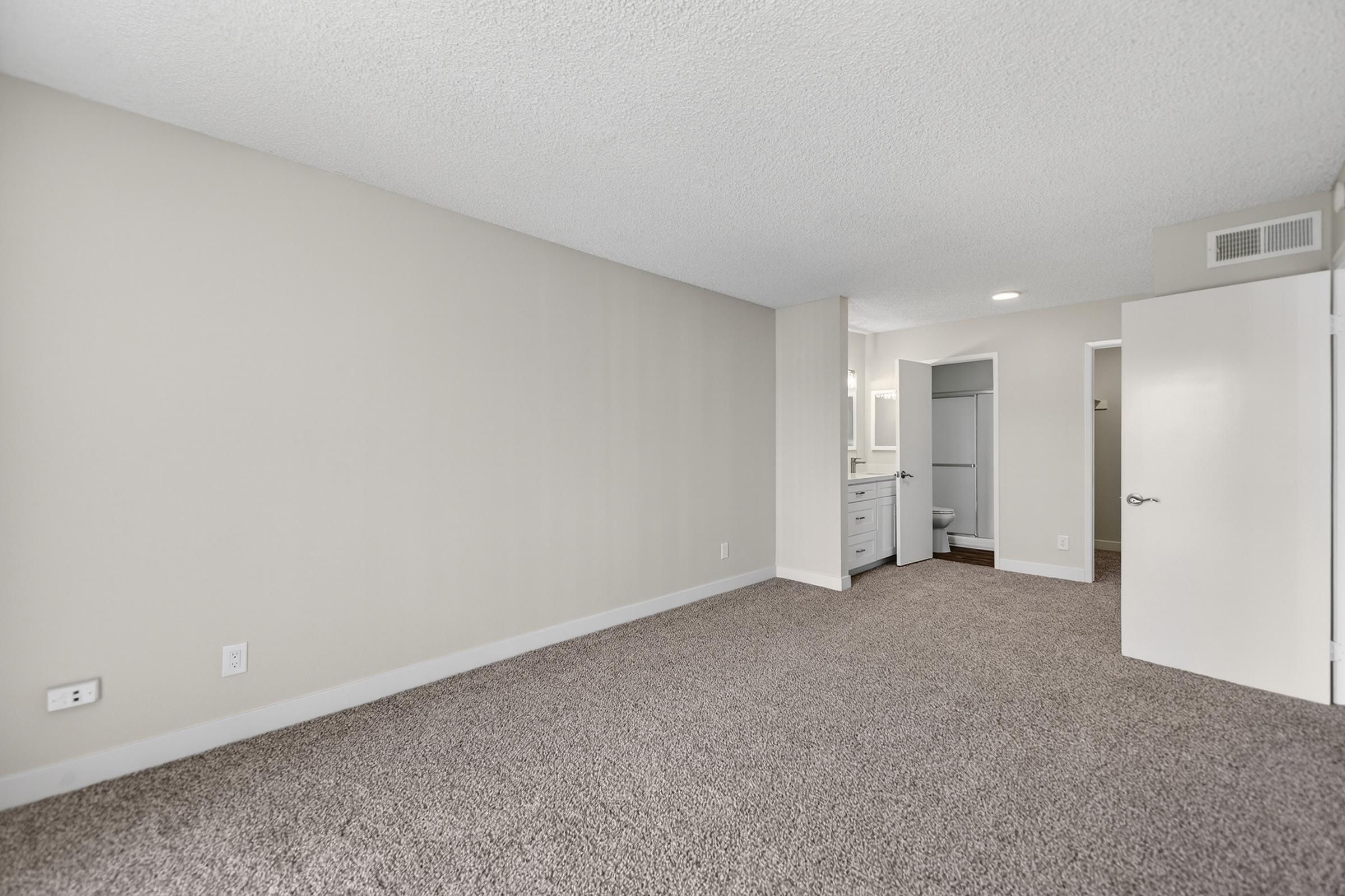 A spacious, neutral-colored room featuring light beige walls and a textured ceiling. The flooring is covered with plush carpet. A doorway leads to a small bathroom area, while another door opens to a closet. Soft natural light brightens the space.